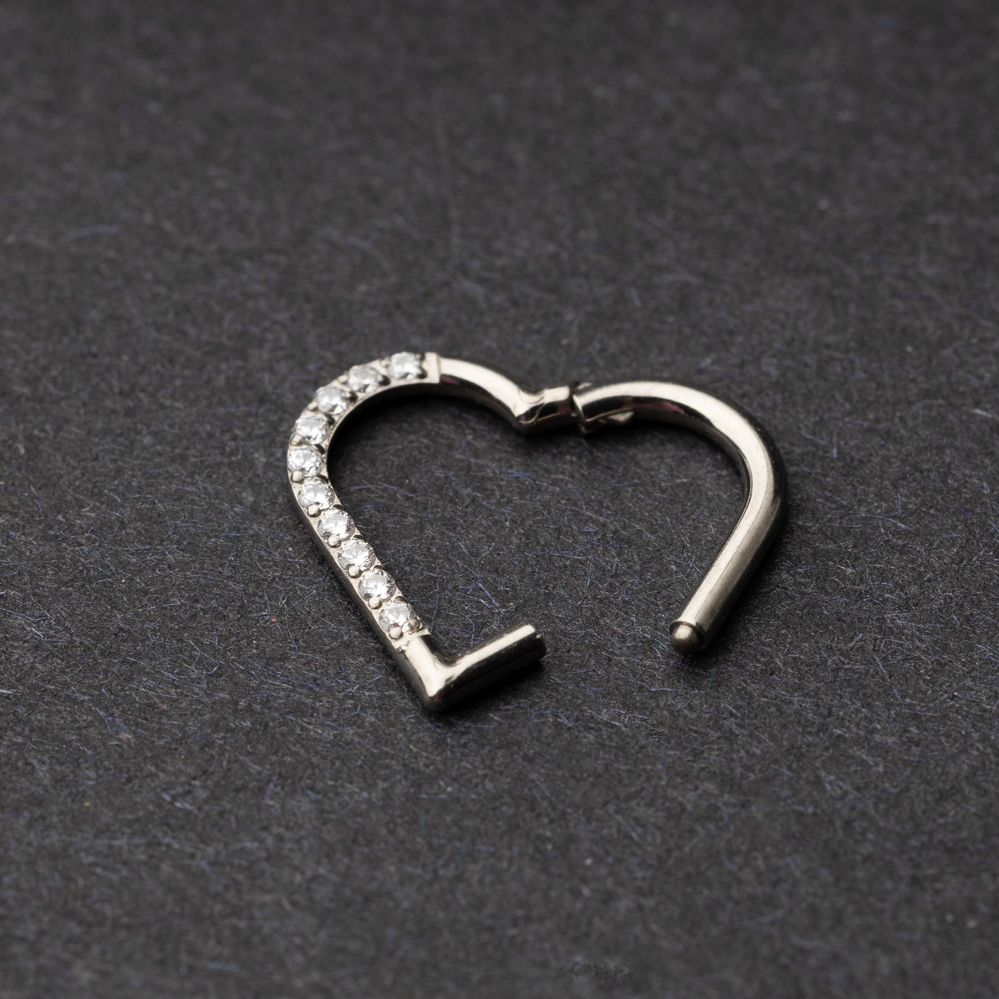 Heart-shaped daith clicker with crystal accents along one side, displayed on a dark textured background.