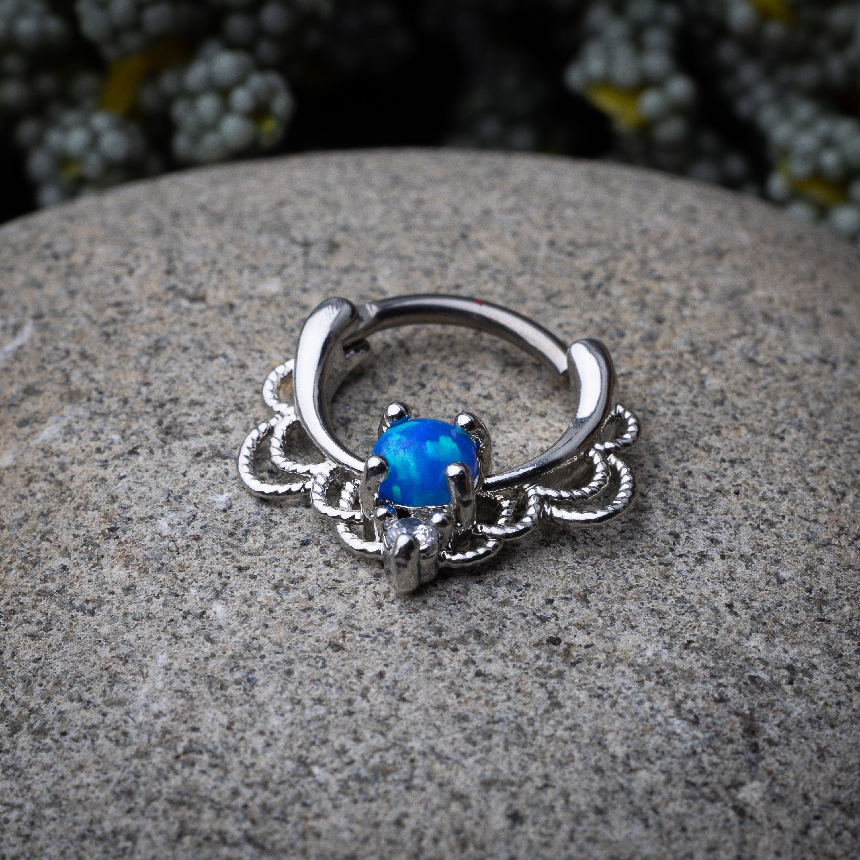 Silver septum clicker with intricate lace design displayed on a stone background.