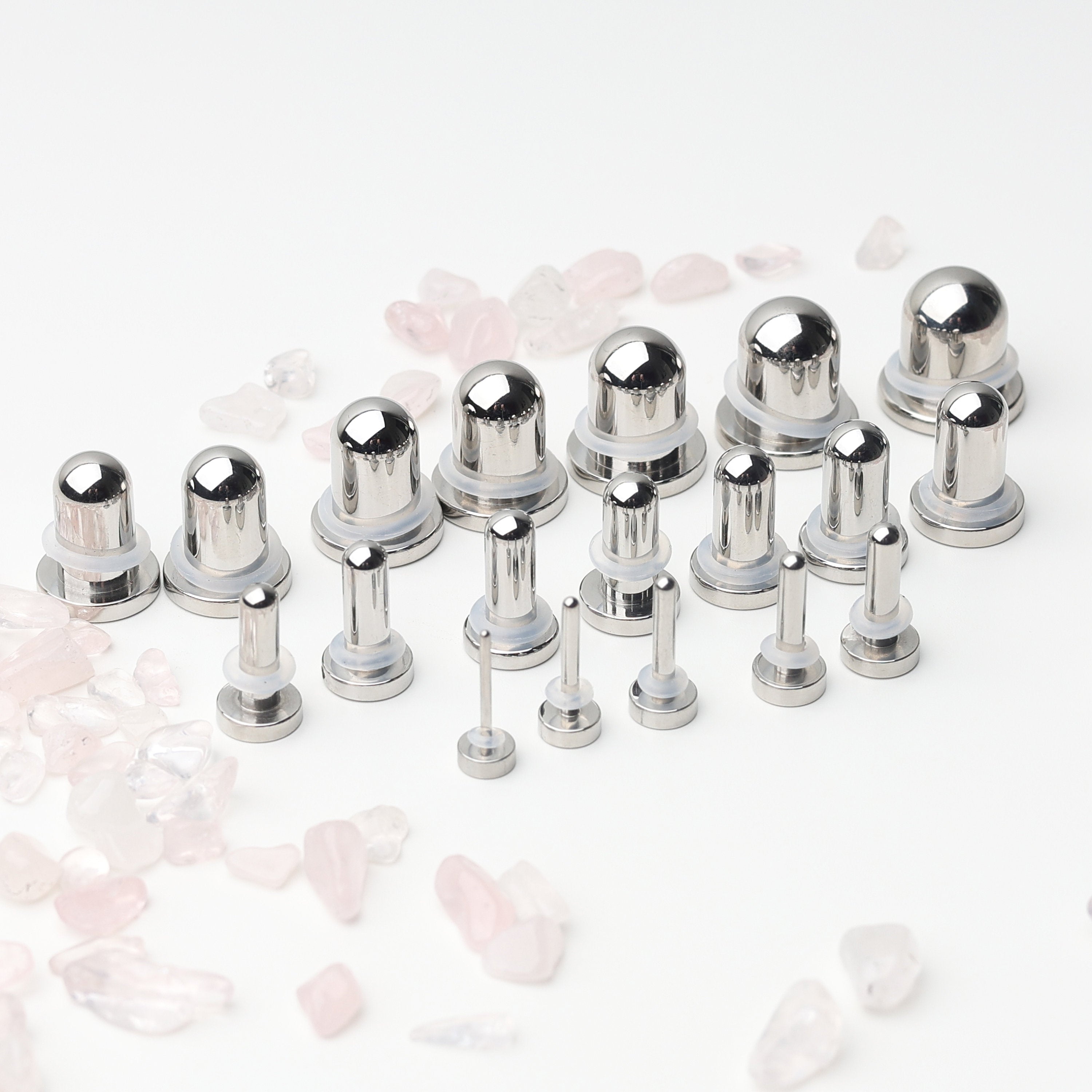 A complete dead stretching kit with polished stainless steel plugs and tunnels in multiple sizes, displayed on a white background with scattered rose quartz accents.