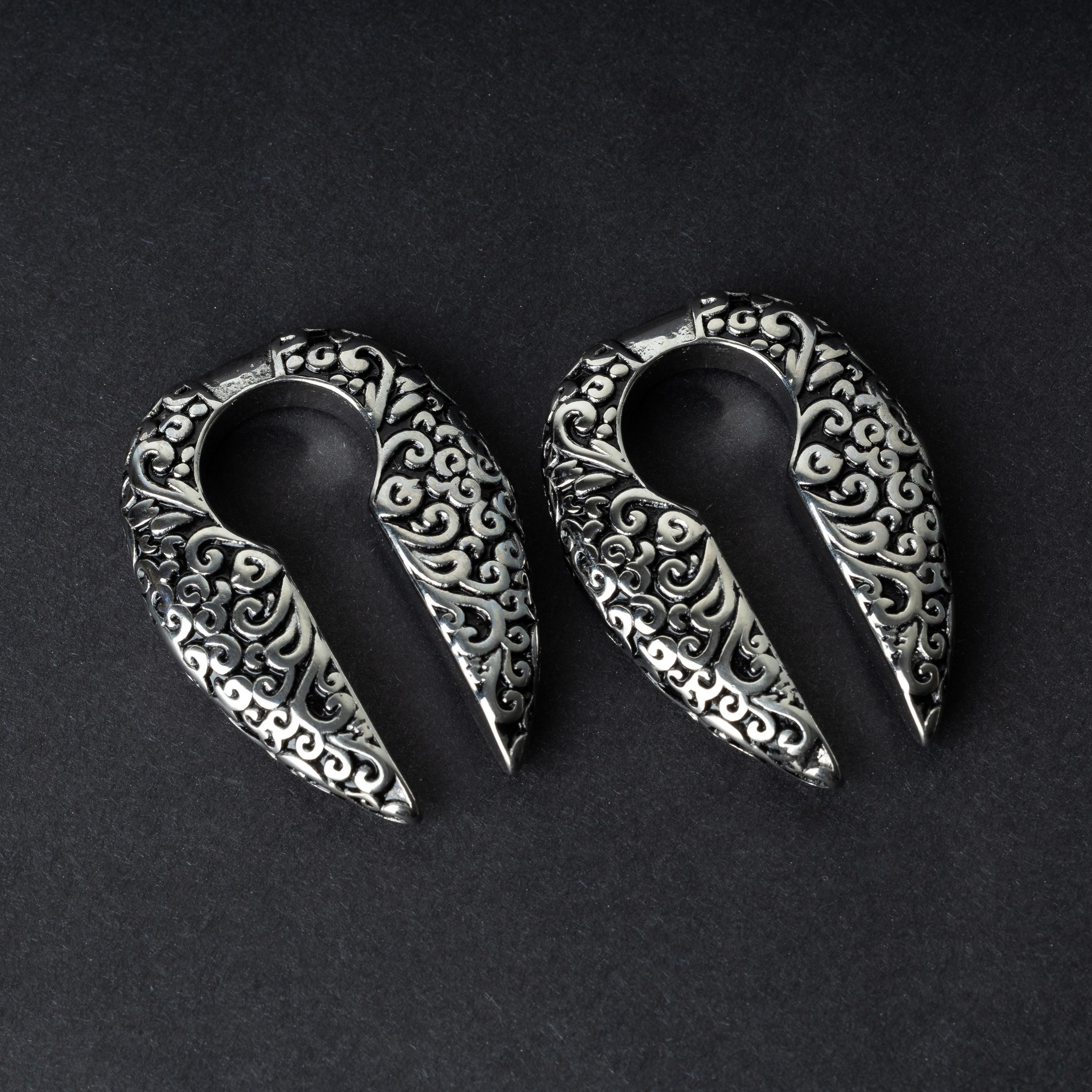 A pair of split ear weights in polished silver with detailed filigree patterns, displayed on a dark background.