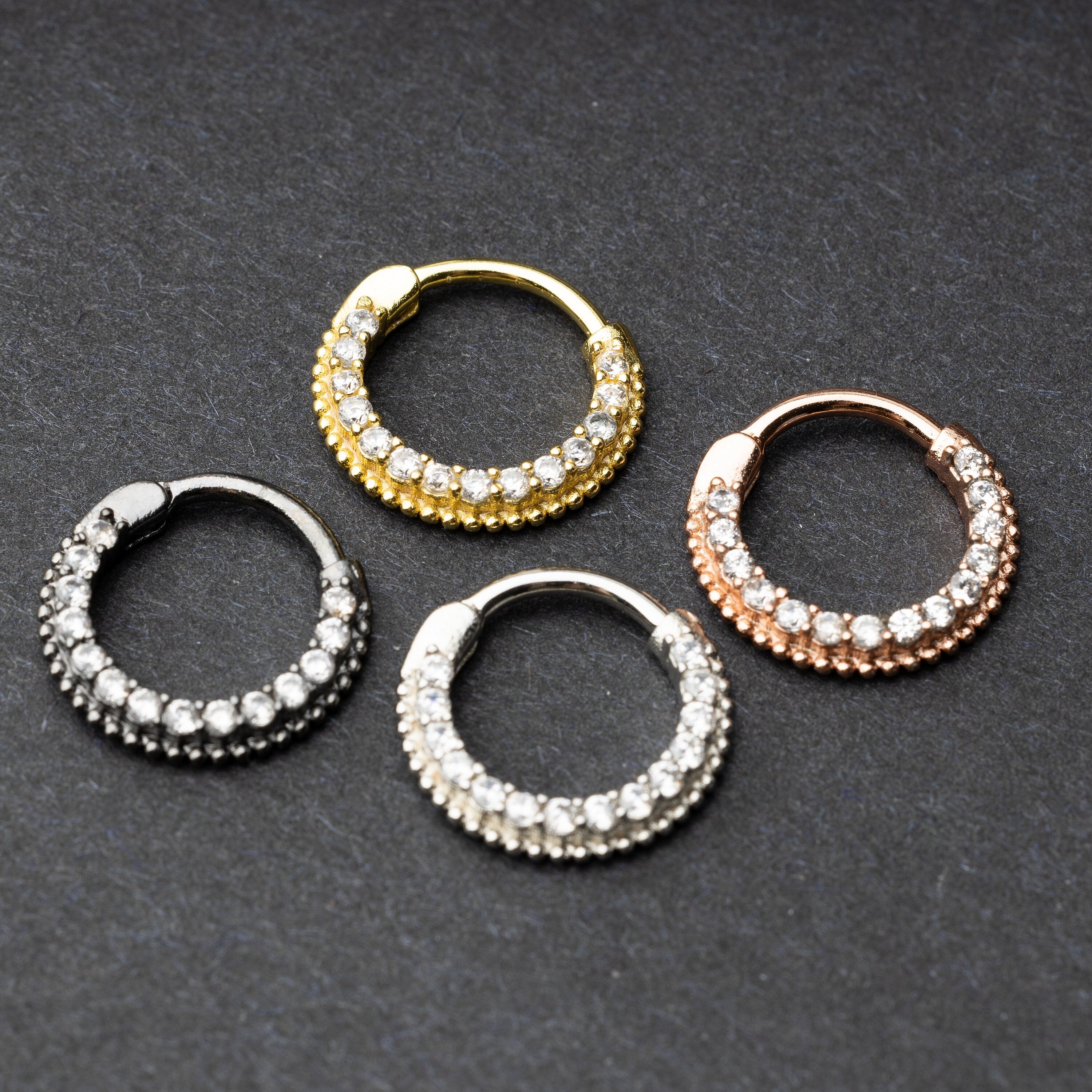 Four stainless steel clickers with double rows of clear gemstones in black, gold, silver, and rose gold finishes, displayed on a dark background