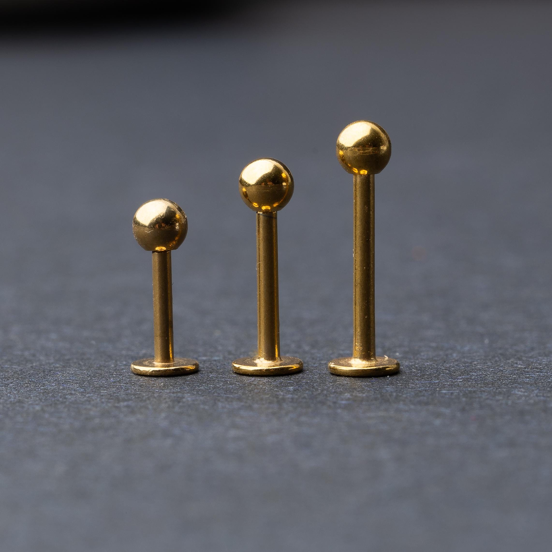 Gold labret studs with ball ends in three different lengths, displayed on a dark background.