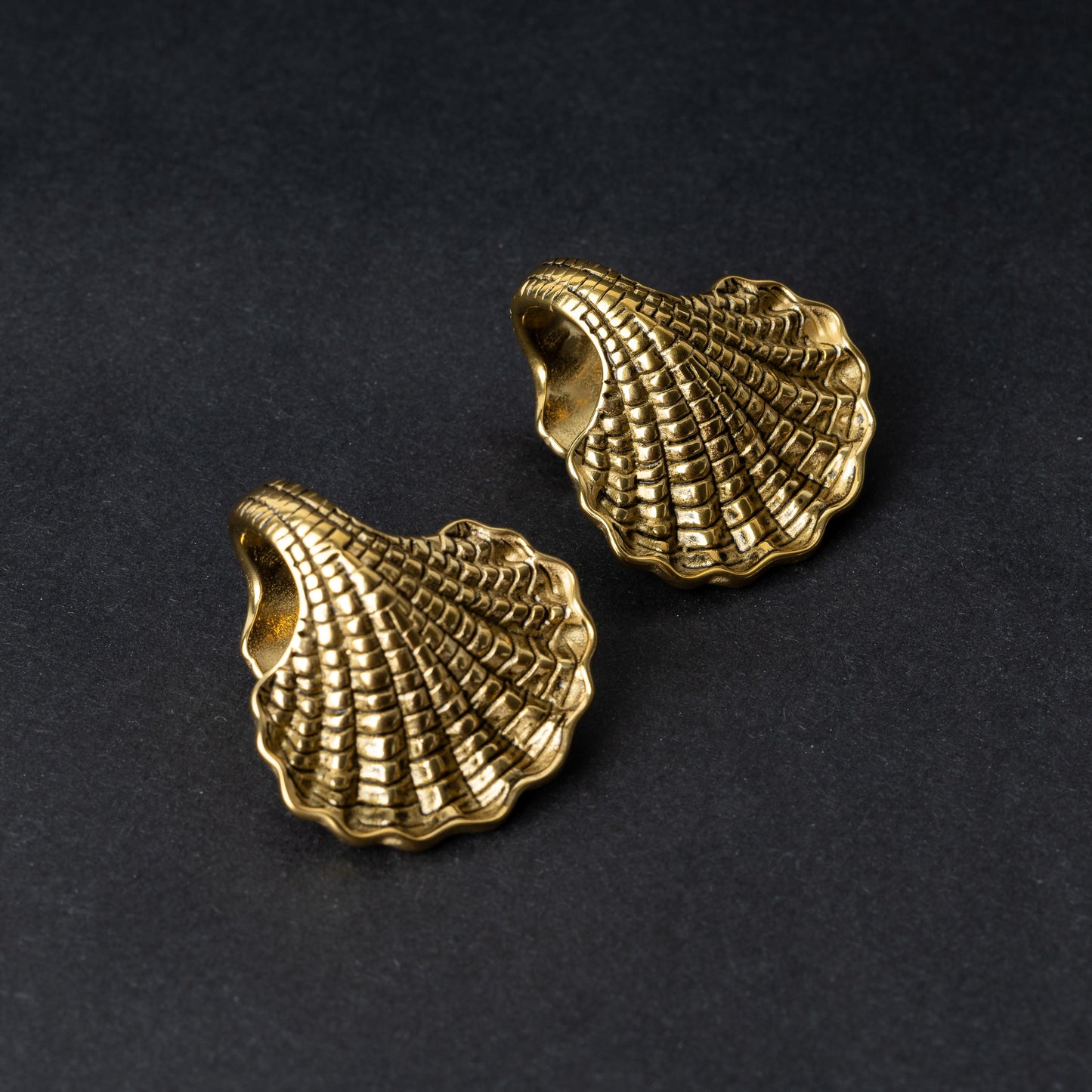 Two seashell-shaped ear weights in polished gold, with detailed textures and a bold design, displayed on a dark background.