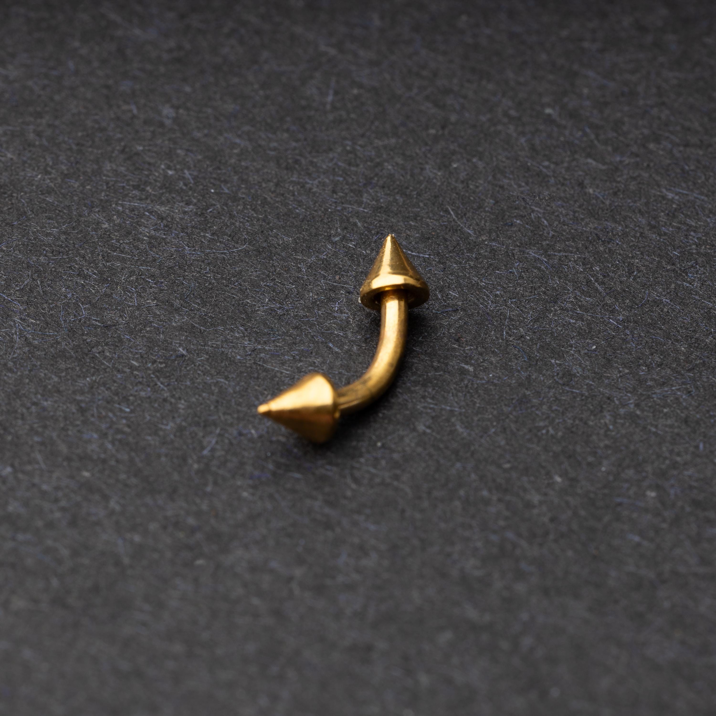 Gold curved barbell with pointed spike ends, displayed on a dark background.