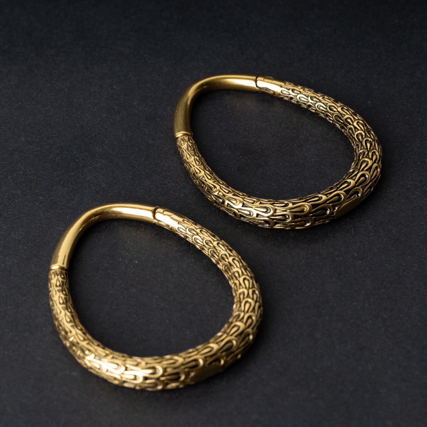 A pair of gold teardrop ear weights featuring a detailed dragon-scale texture and a seamless hinge closure, displayed on a dark background.
