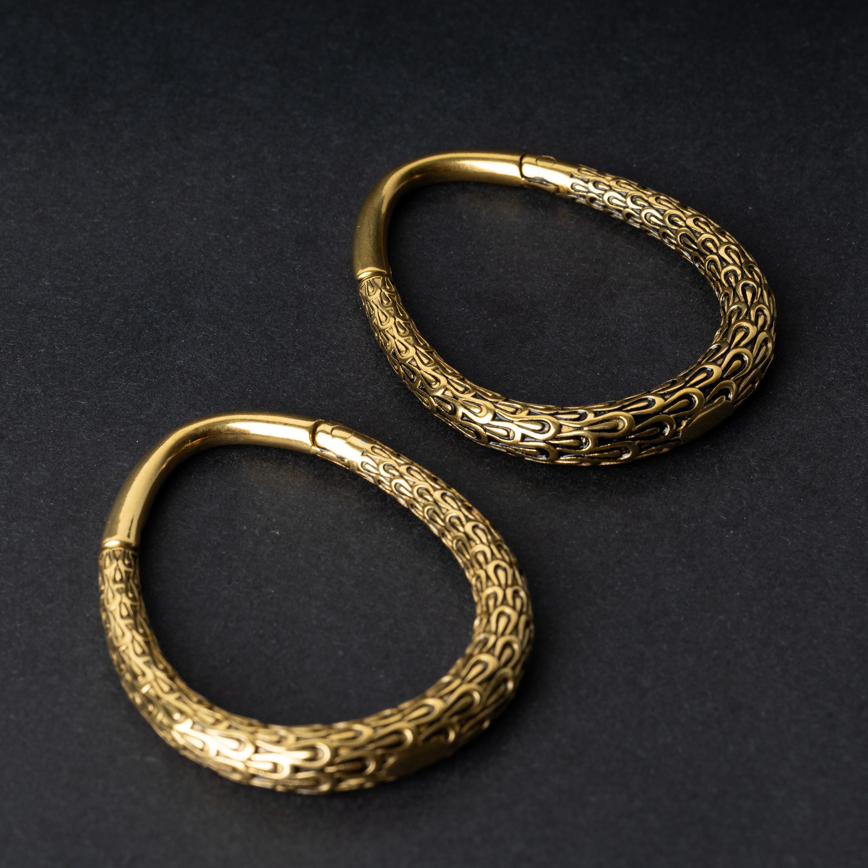 A pair of gold teardrop ear weights featuring a detailed dragon-scale texture and a seamless hinge closure, displayed on a dark background.