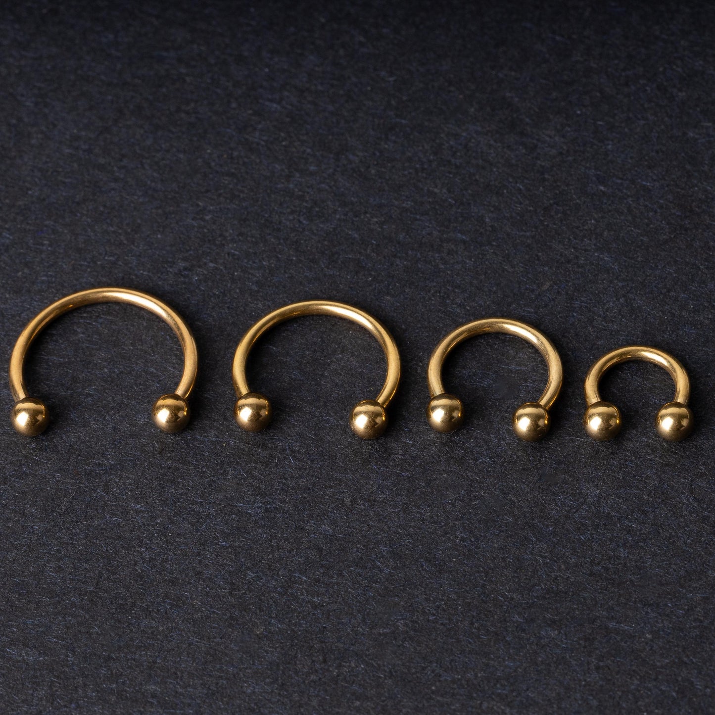 Set of four gold-colored titanium horseshoe rings with ball ends, displayed on a dark textured background.