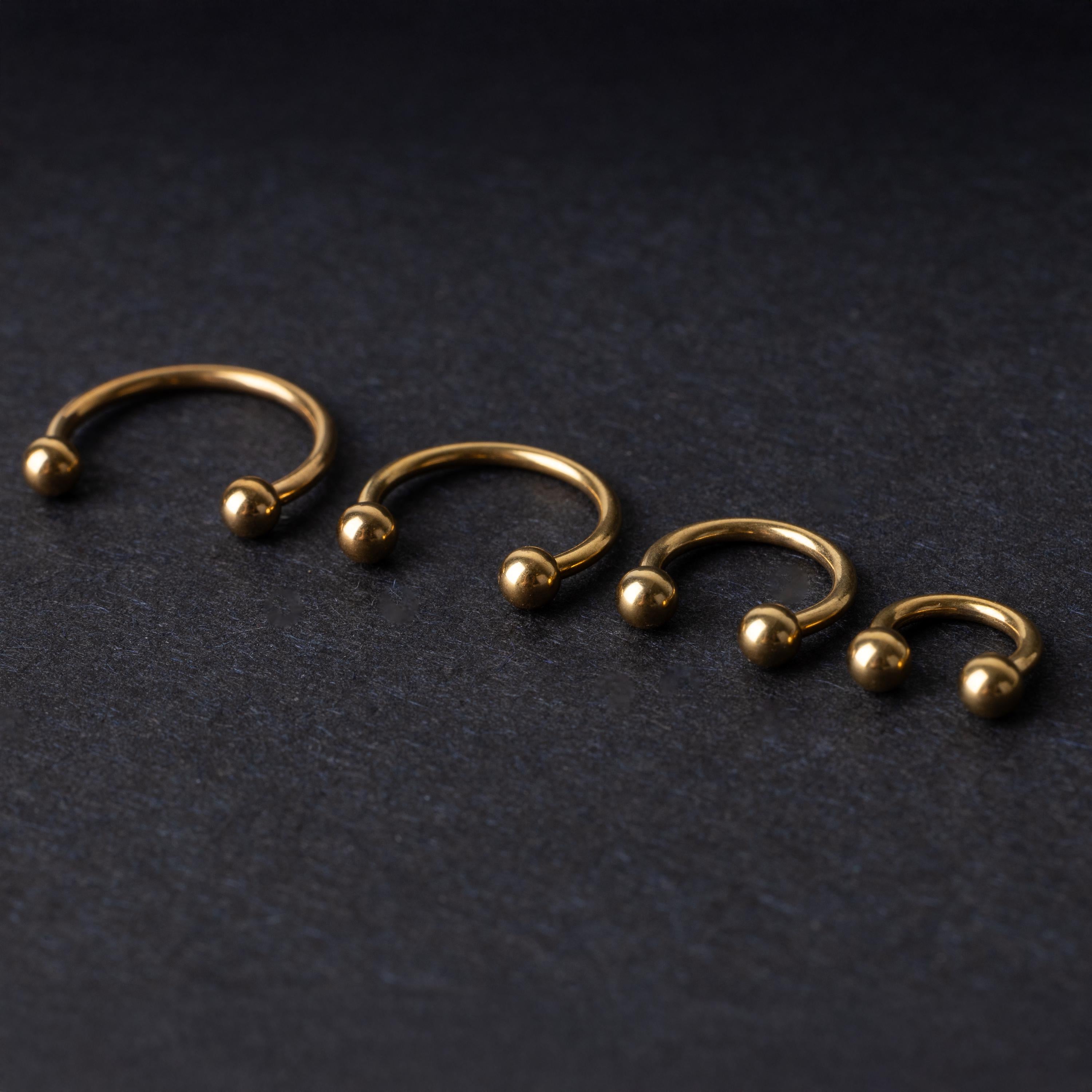 Set of four gold-colored titanium horseshoe rings with ball ends, displayed on a dark textured background.
