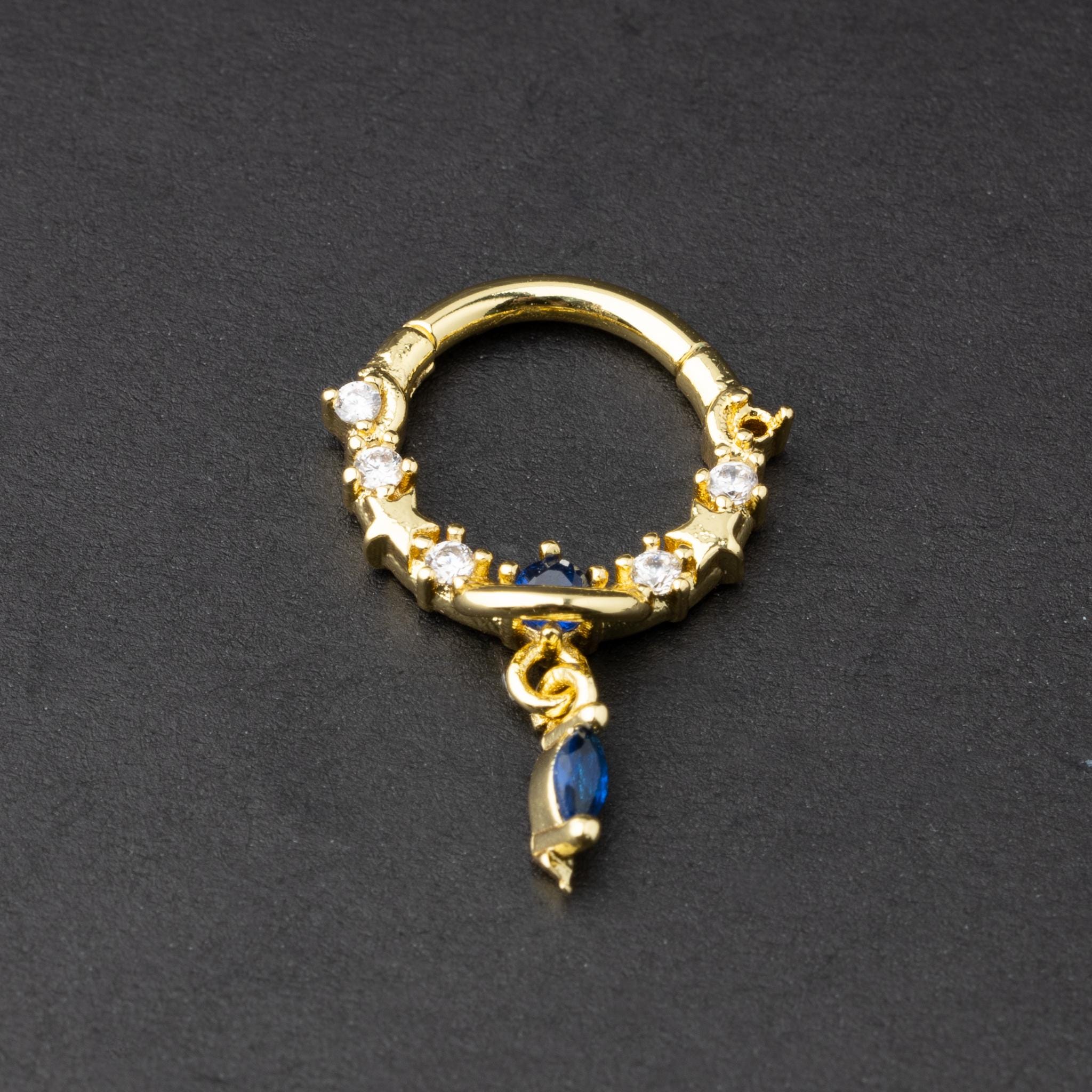 Gold and silver septum clickers adorned with cubic zirconia and a dangling blue gemstone charm, displayed on a dark background.