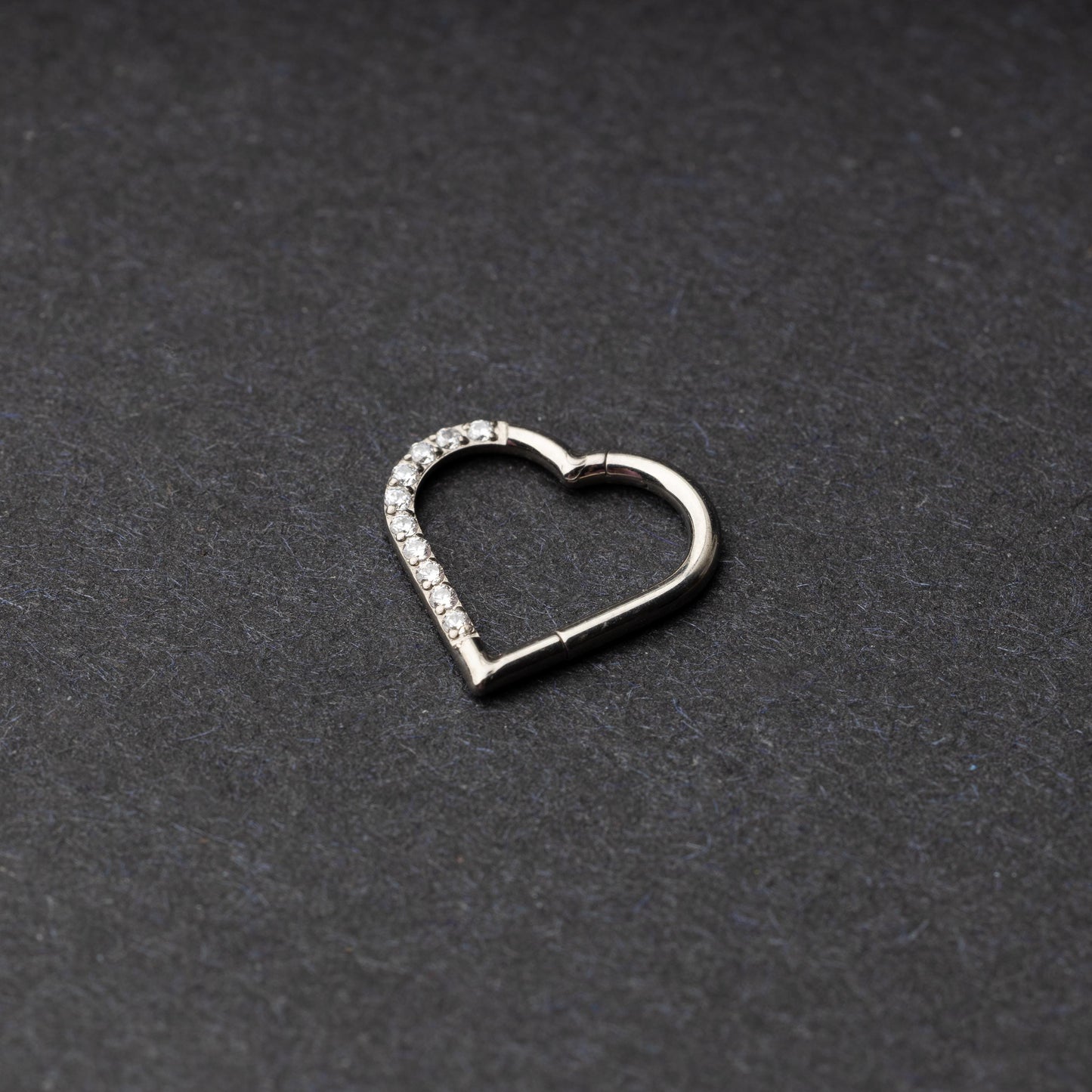 Heart-shaped daith clicker with crystal accents along one side, displayed on a dark textured background.