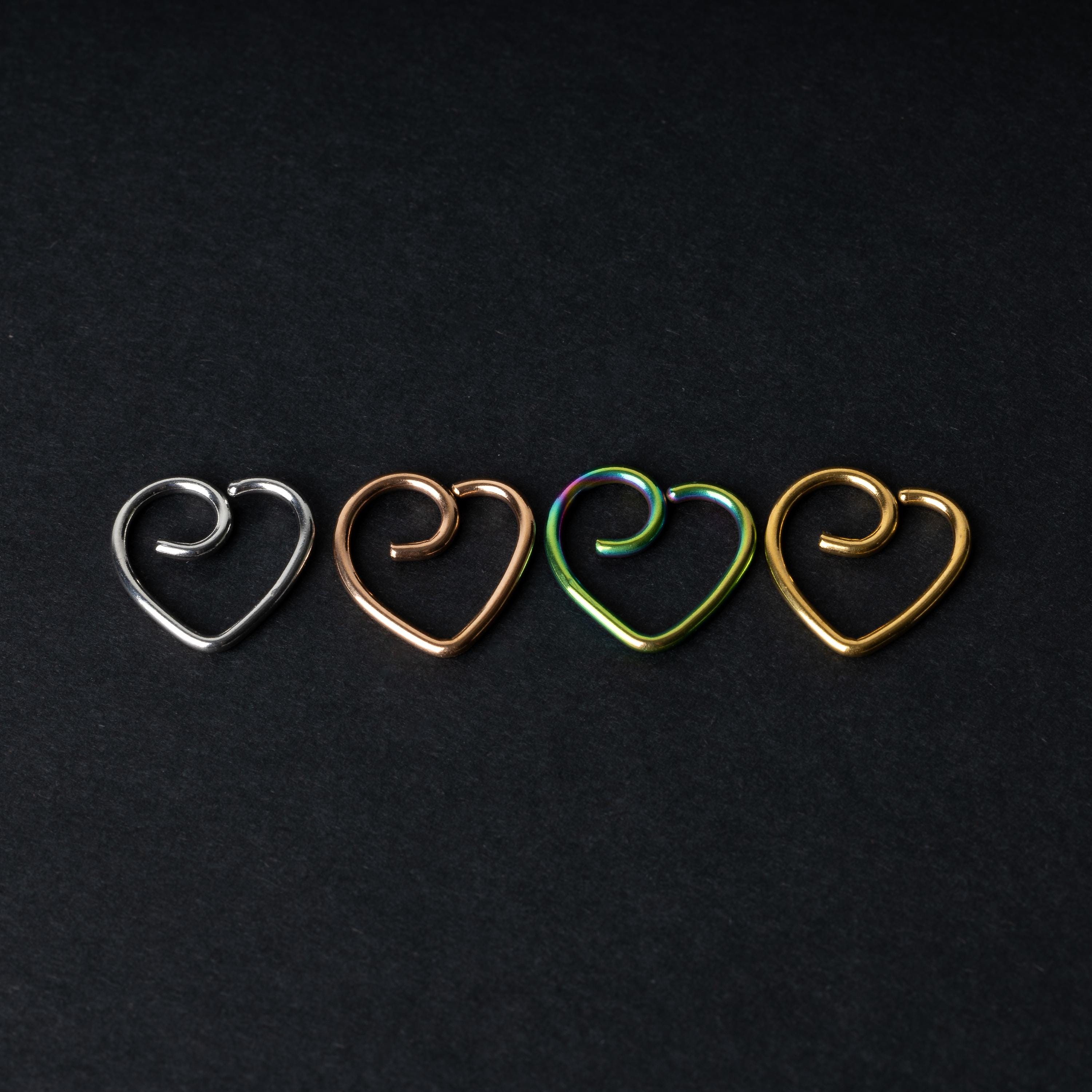 Four heart-shaped nose rings in silver, rose gold, rainbow, and gold finishes, displayed on a dark background.