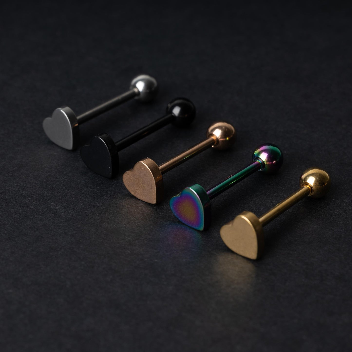 Heart-shaped tongue bars in black, silver, rose gold, rainbow, and gold finishes, displayed on a dark background.