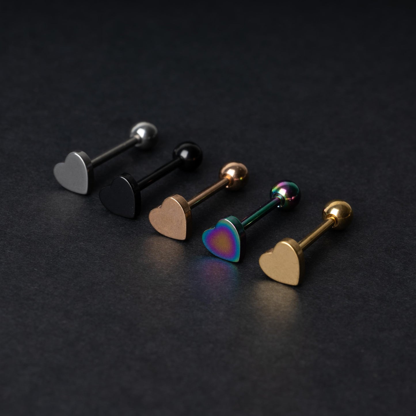 Heart-shaped tongue bars in black, silver, rose gold, rainbow, and gold finishes, displayed on a dark background.