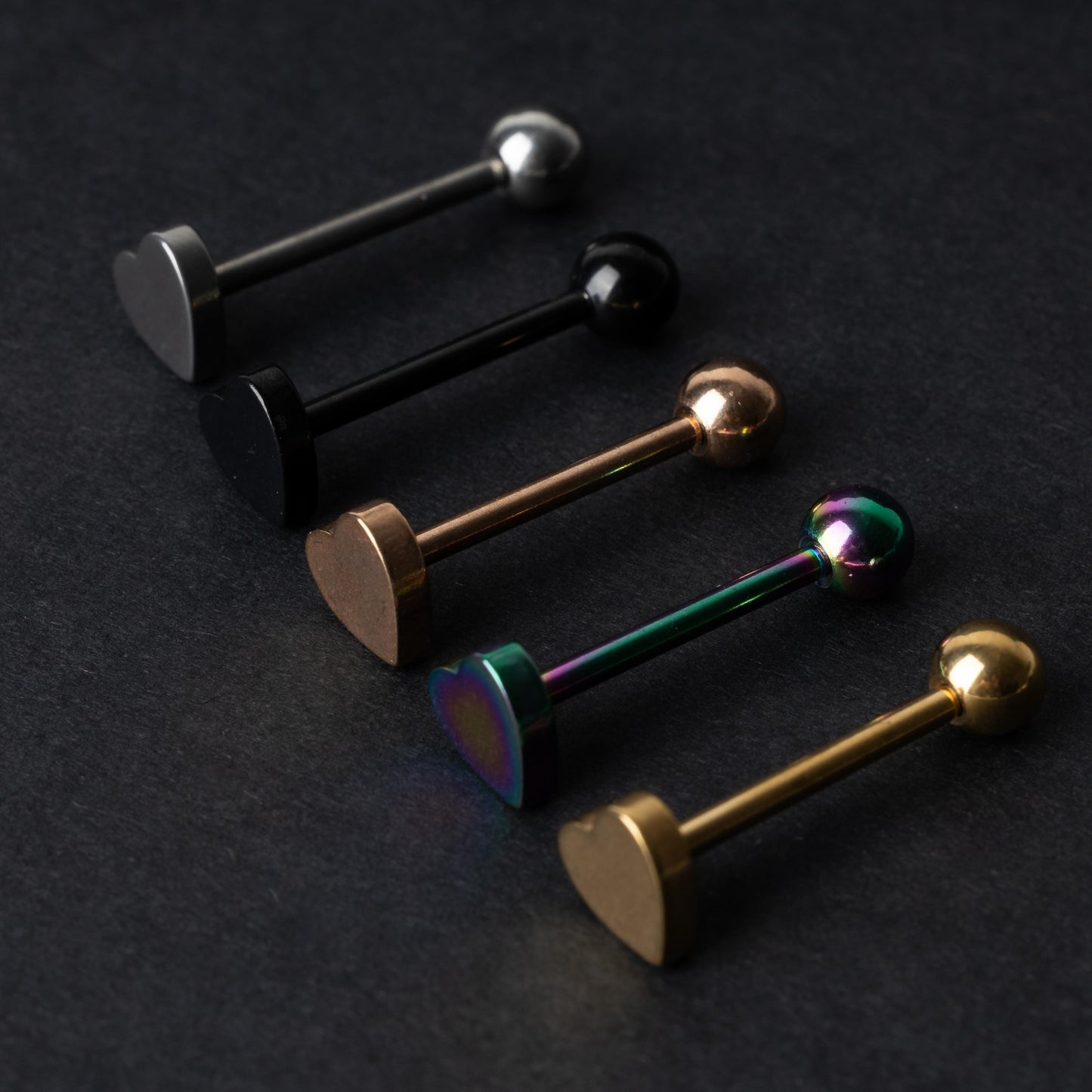 Heart-shaped tongue bars in black, silver, rose gold, rainbow, and gold finishes, displayed on a dark background.