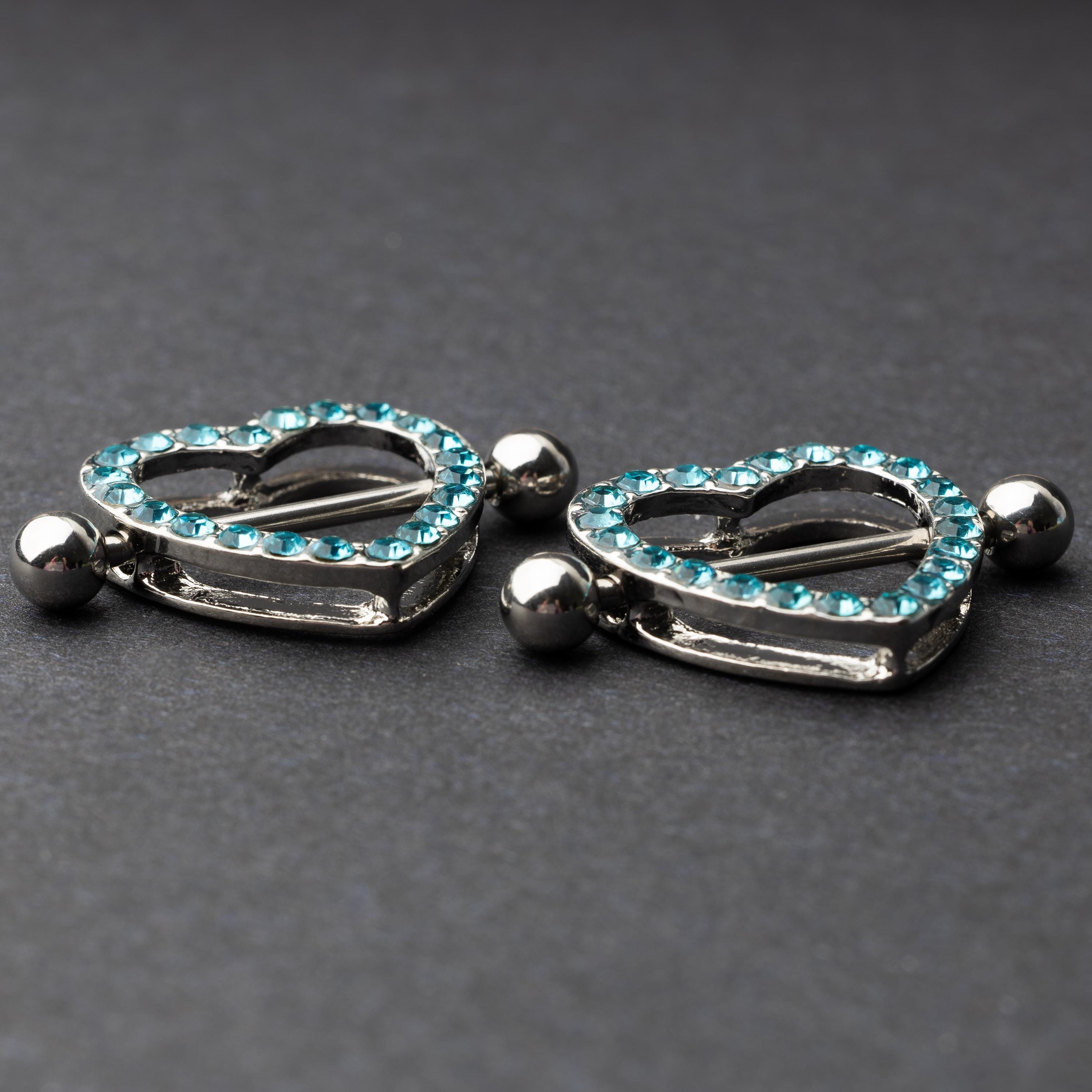 Two heart-shaped nipple barbells with silver metal and light blue gemstone accents, displayed on a dark background.