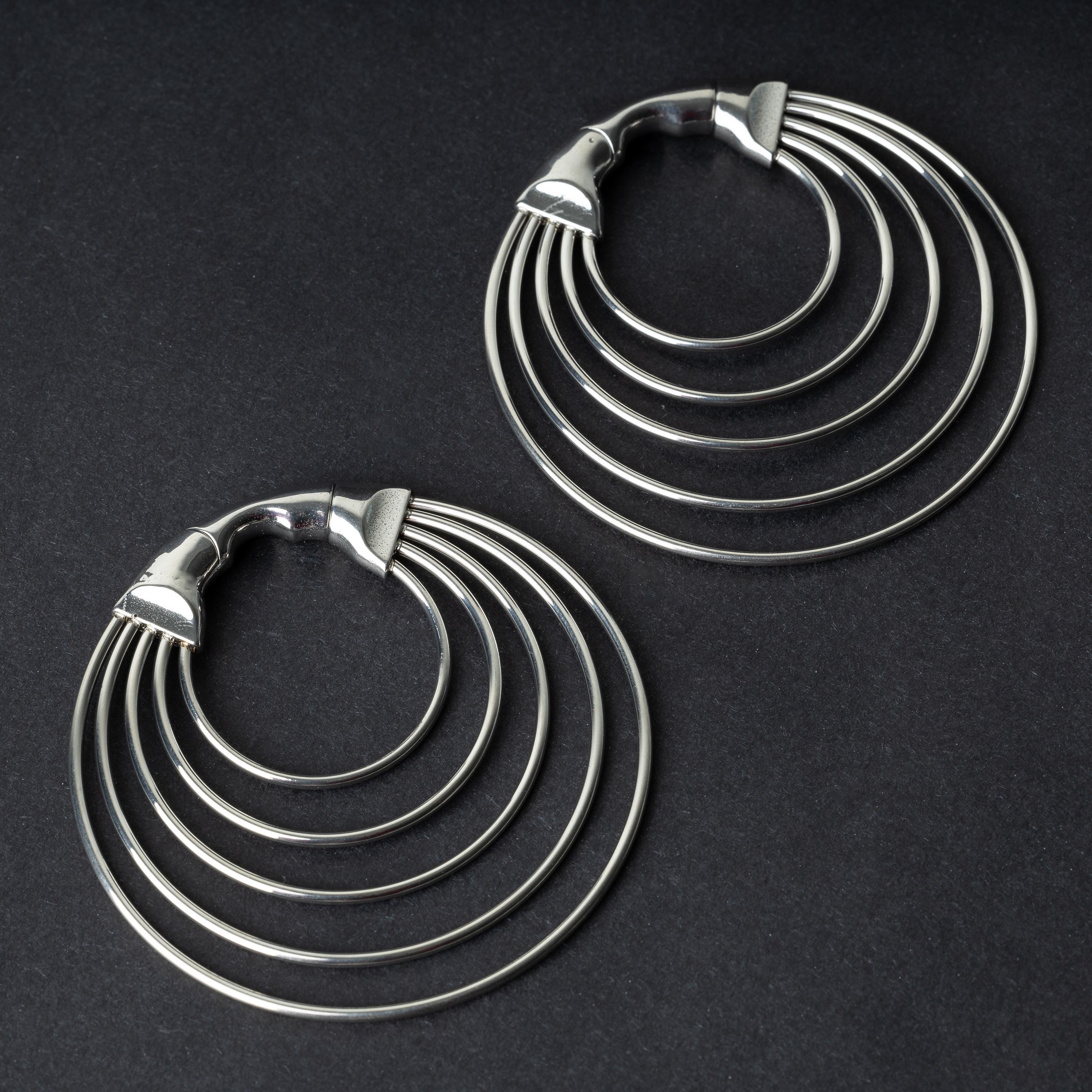 A pair of multi-hoop ear weights in polished silver, featuring five cascading hoops and a seamless hinge closure, displayed on a dark background.