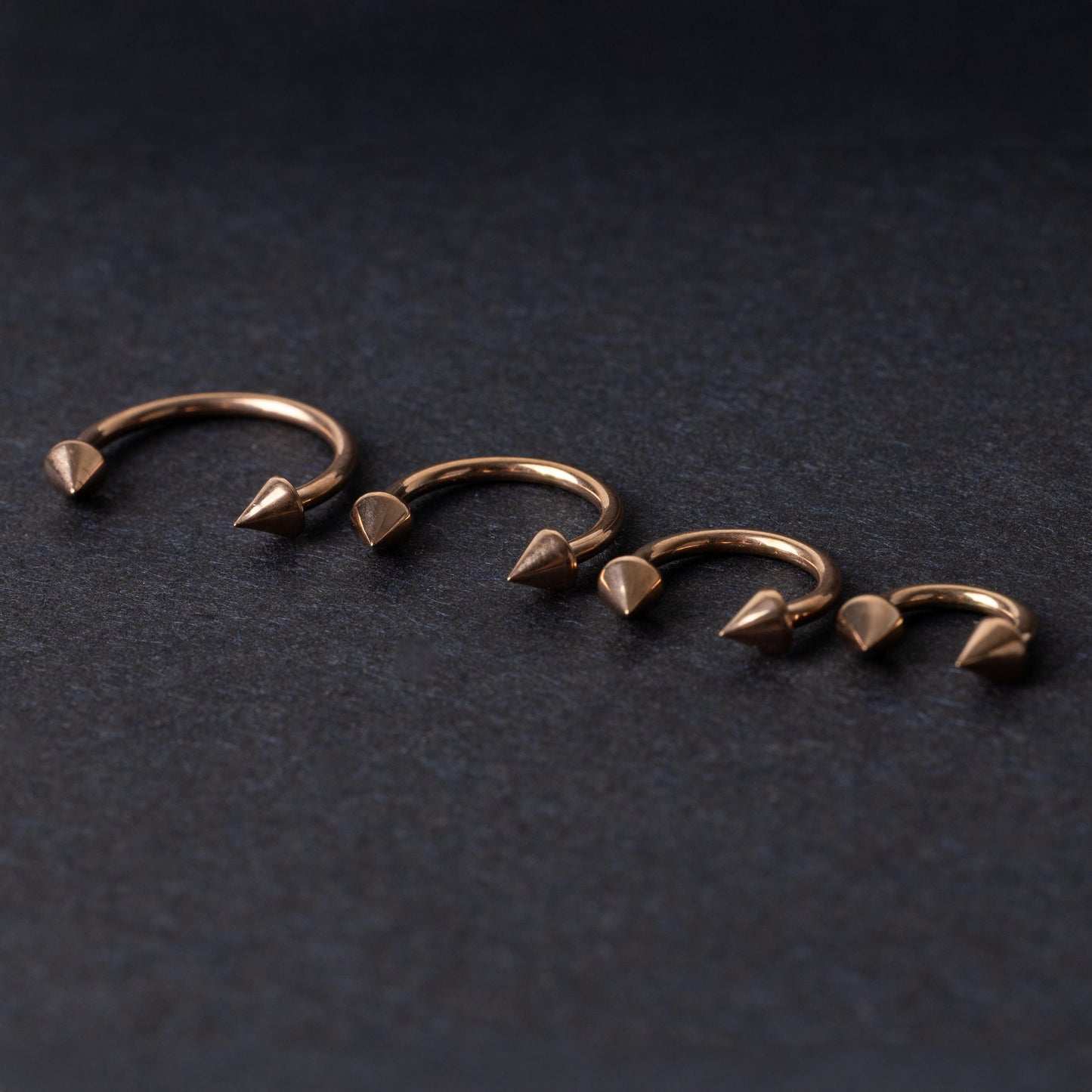 Four rose gold-tone horseshoe hoops with spike ends, arranged in descending size order on a dark textured background.