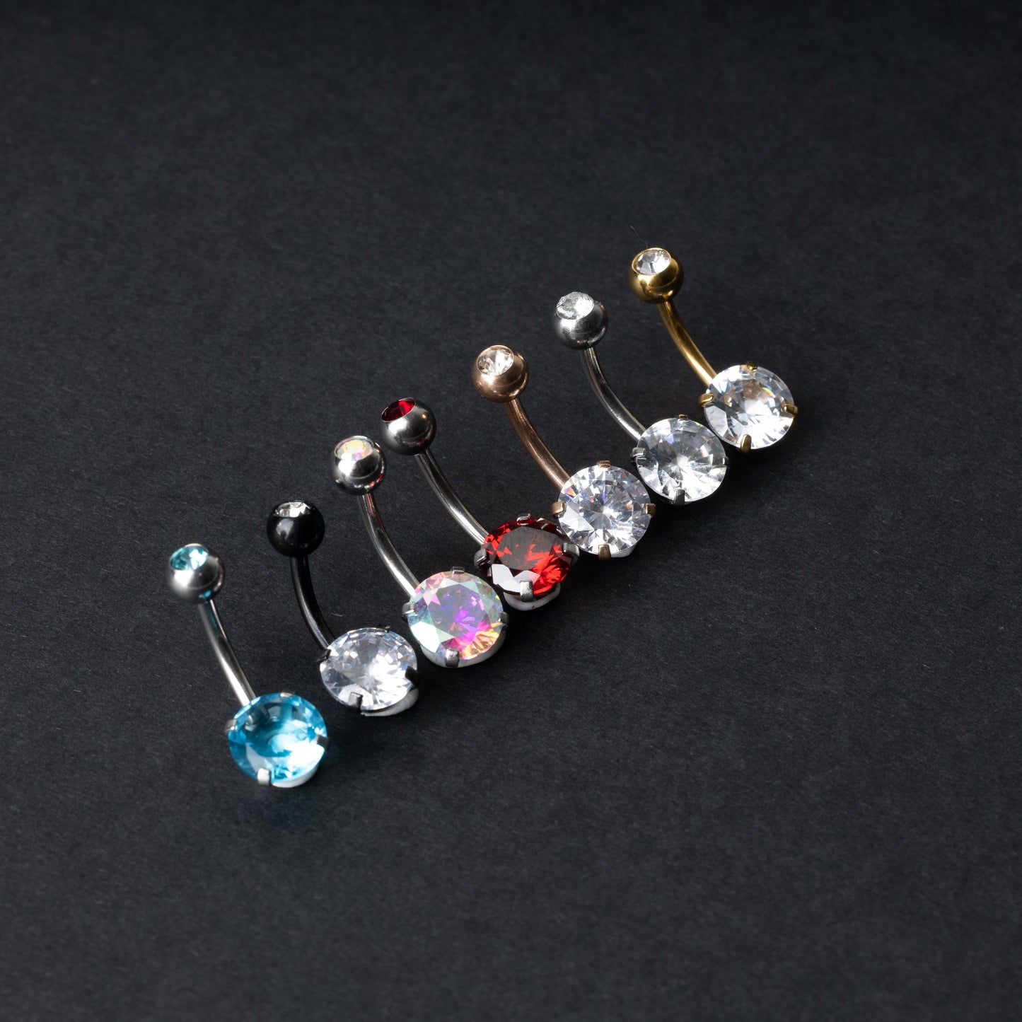 A collection of colorful belly button rings with curved barbells, featuring large gemstone centerpieces in shades of blue, black, clear, iridescent, red, and gold, displayed on a black background.