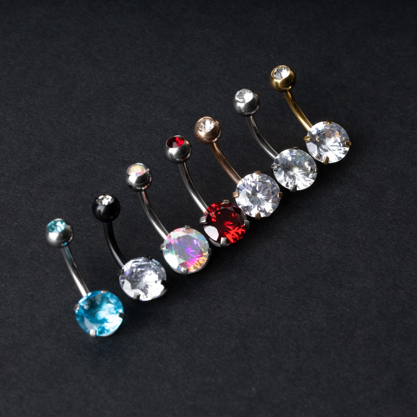 A collection of colorful belly button rings with curved barbells, featuring large gemstone centerpieces in shades of blue, black, clear, iridescent, red, and gold, displayed on a black background.