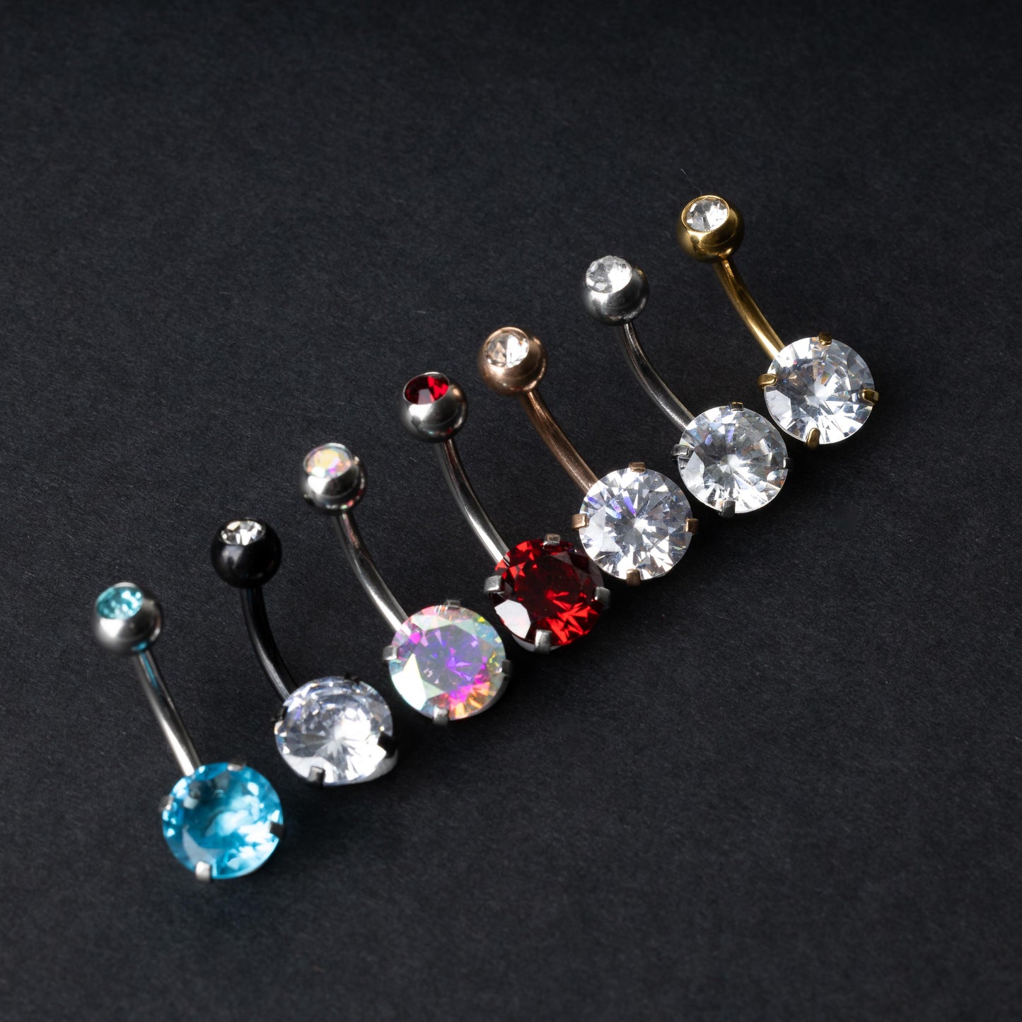 A collection of colorful belly button rings with curved barbells, featuring large gemstone centerpieces in shades of blue, black, clear, iridescent, red, and gold, displayed on a black background.