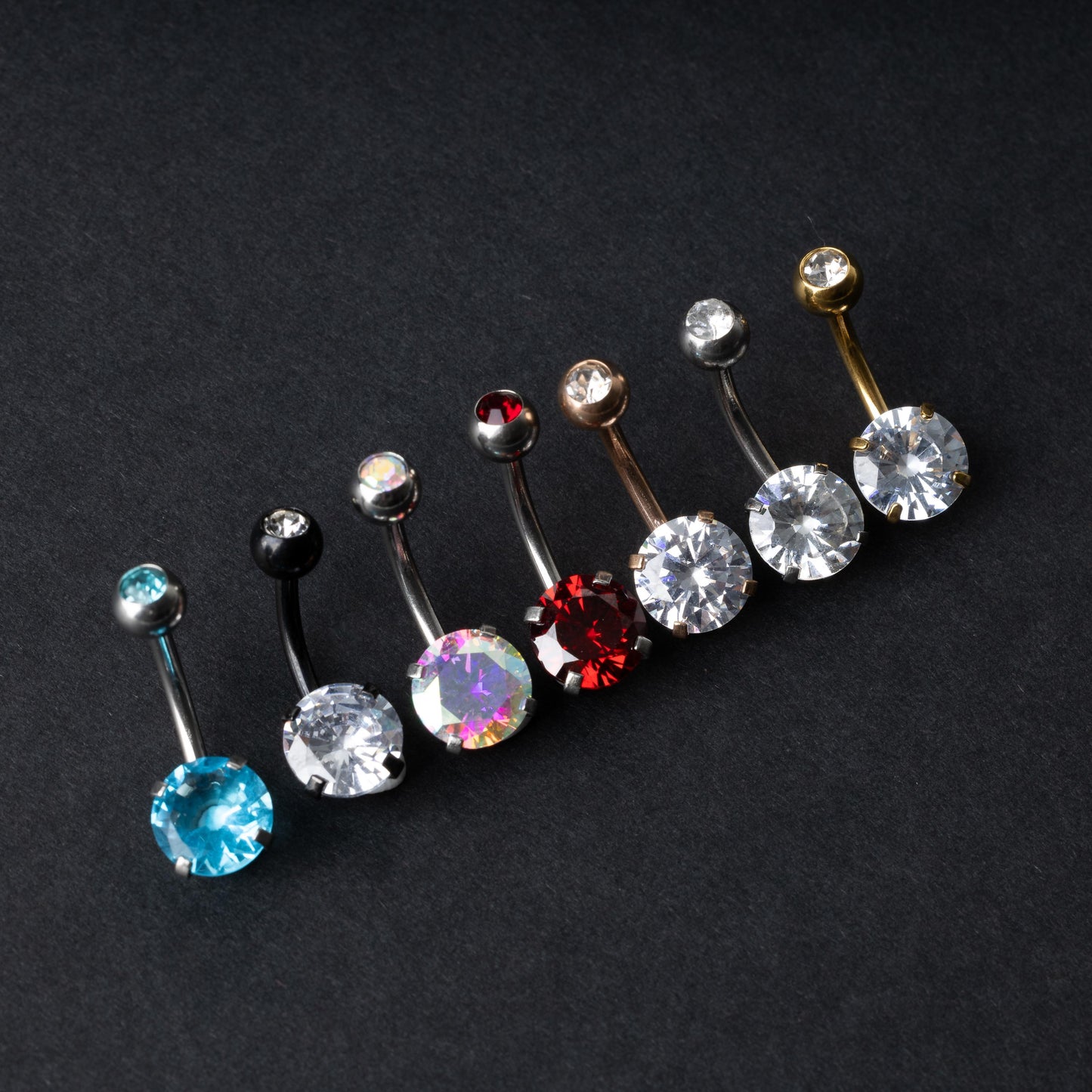 A collection of colorful belly button rings with curved barbells, featuring large gemstone centerpieces in shades of blue, black, clear, iridescent, red, and gold, displayed on a black background.