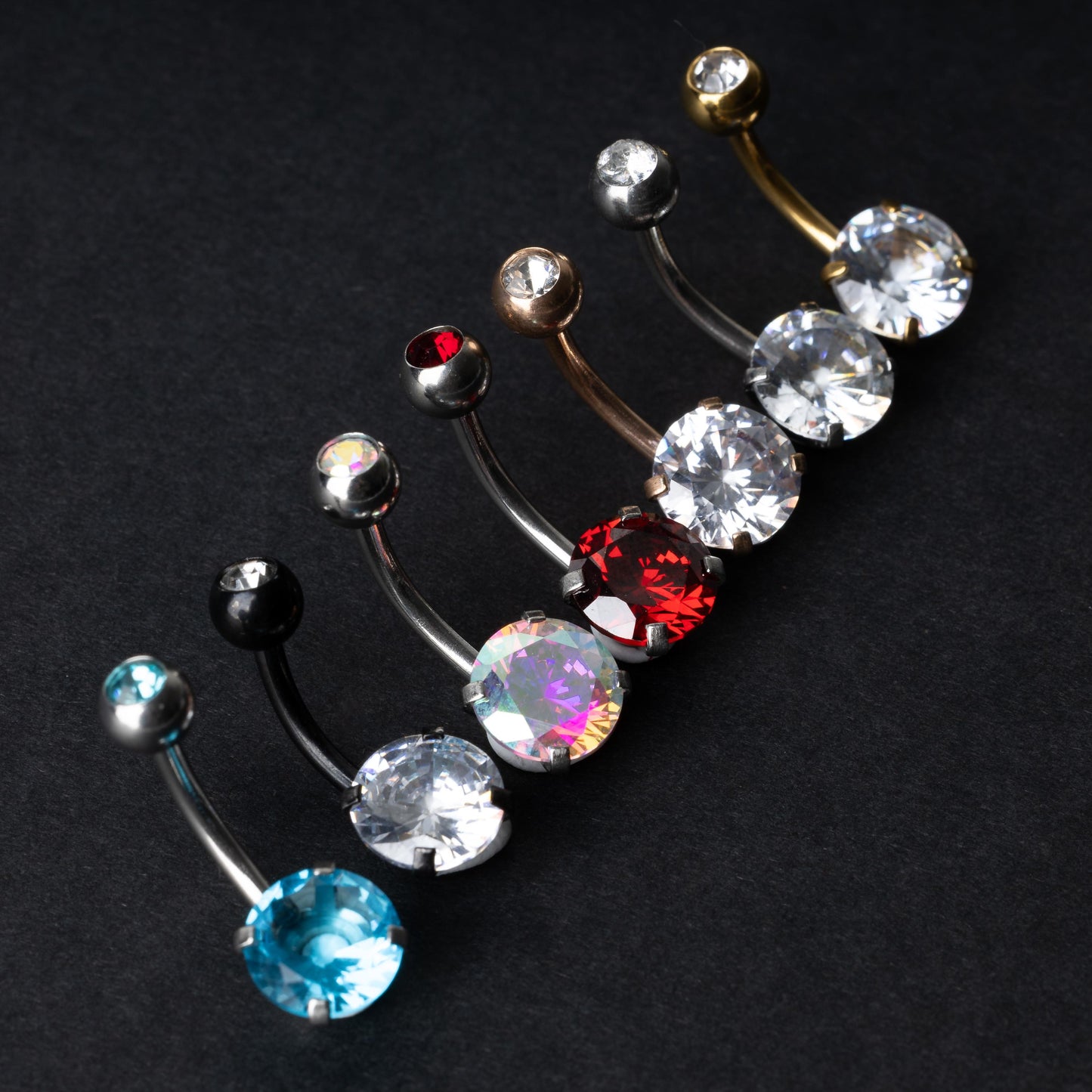 A collection of colorful belly button rings with curved barbells, featuring large gemstone centerpieces in shades of blue, black, clear, iridescent, red, and gold, displayed on a black background.