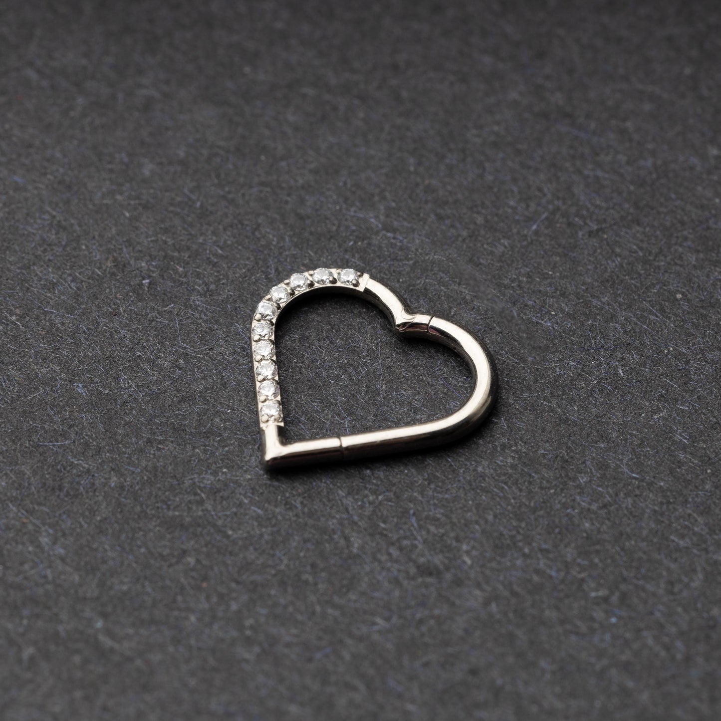 Heart-shaped daith clicker with crystal accents along one side, displayed on a dark textured background.