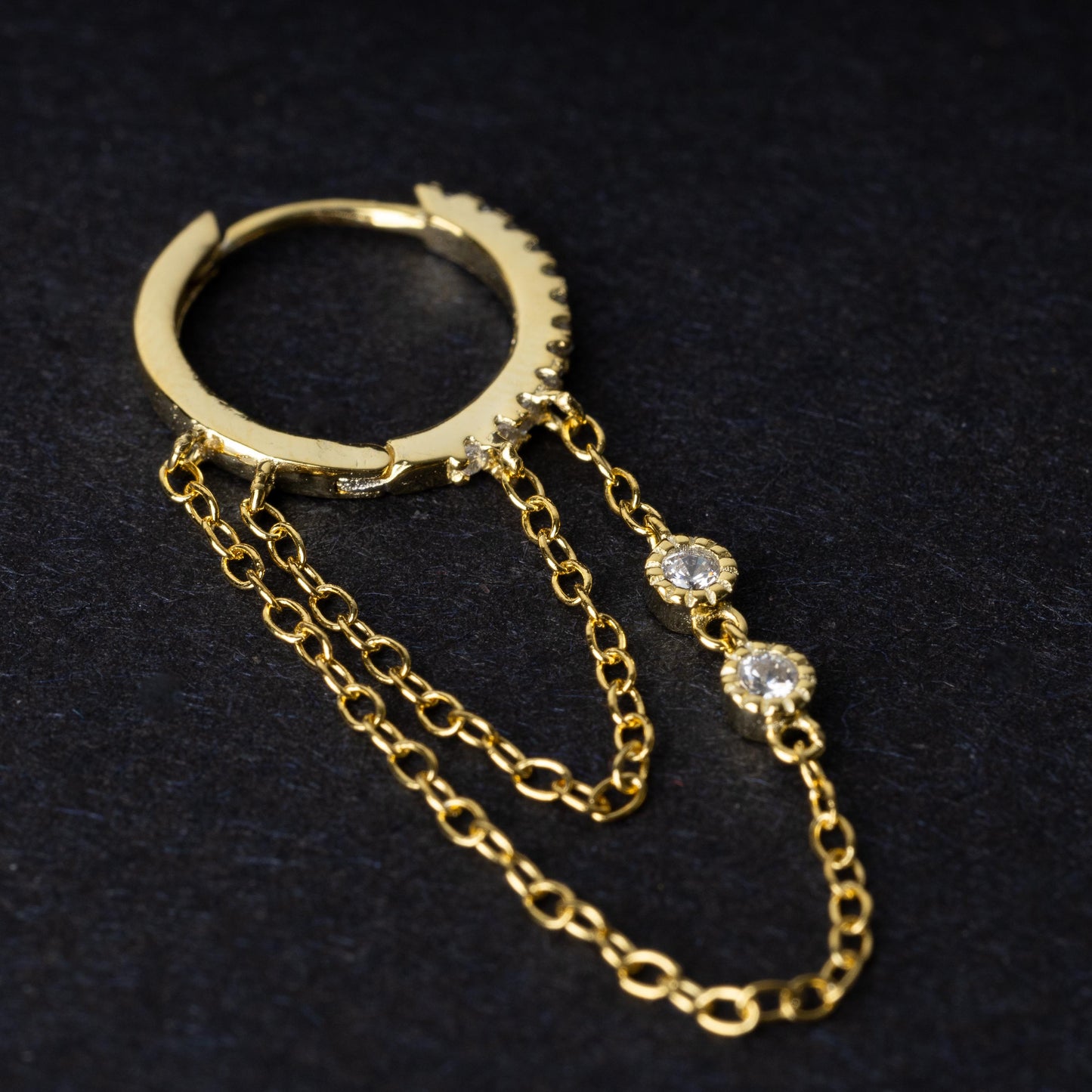 A gold septum clicker with a decorative design featuring dual chains and cubic zirconia gems, displayed on a black background.