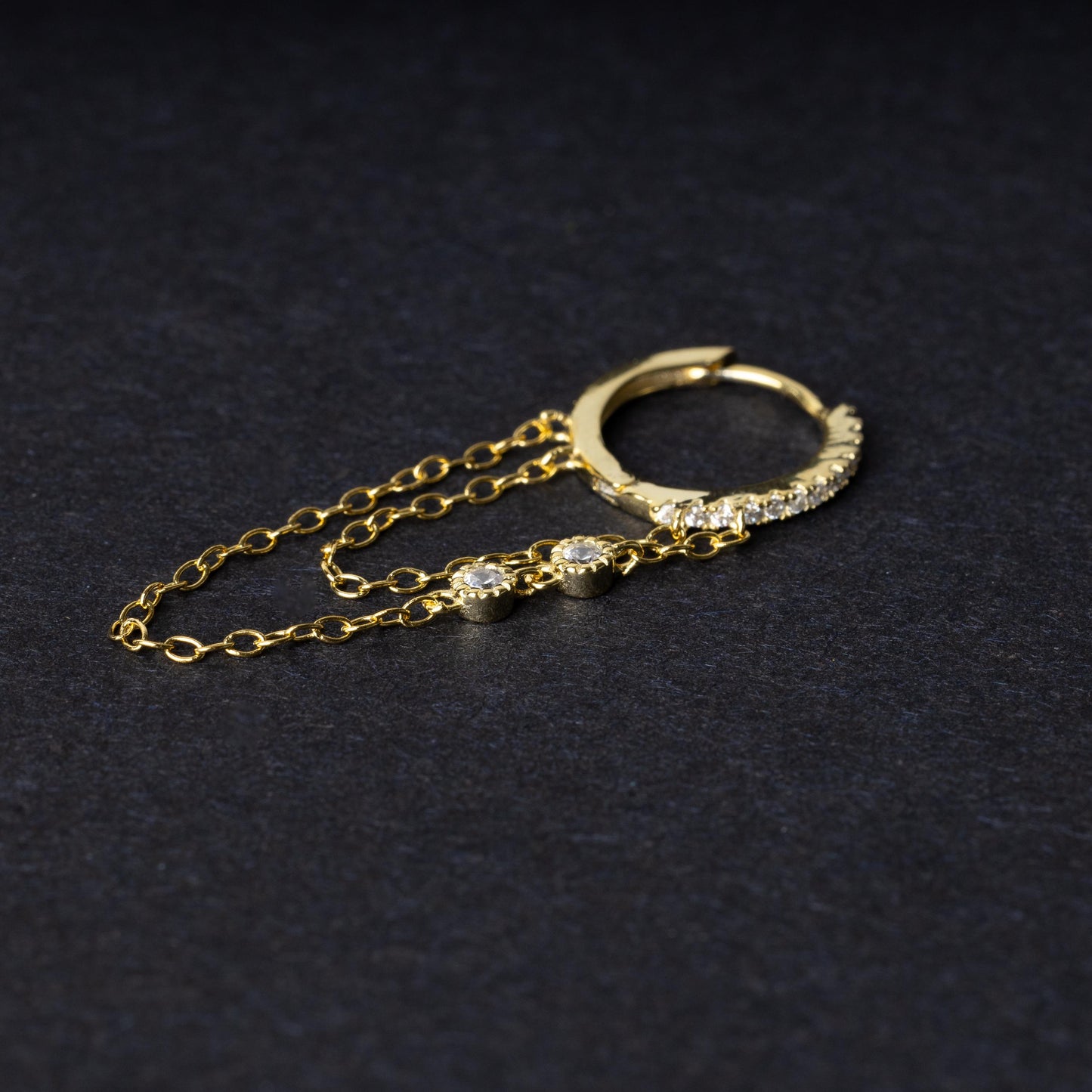 A gold septum clicker with a decorative design featuring dual chains and cubic zirconia gems, displayed on a black background.