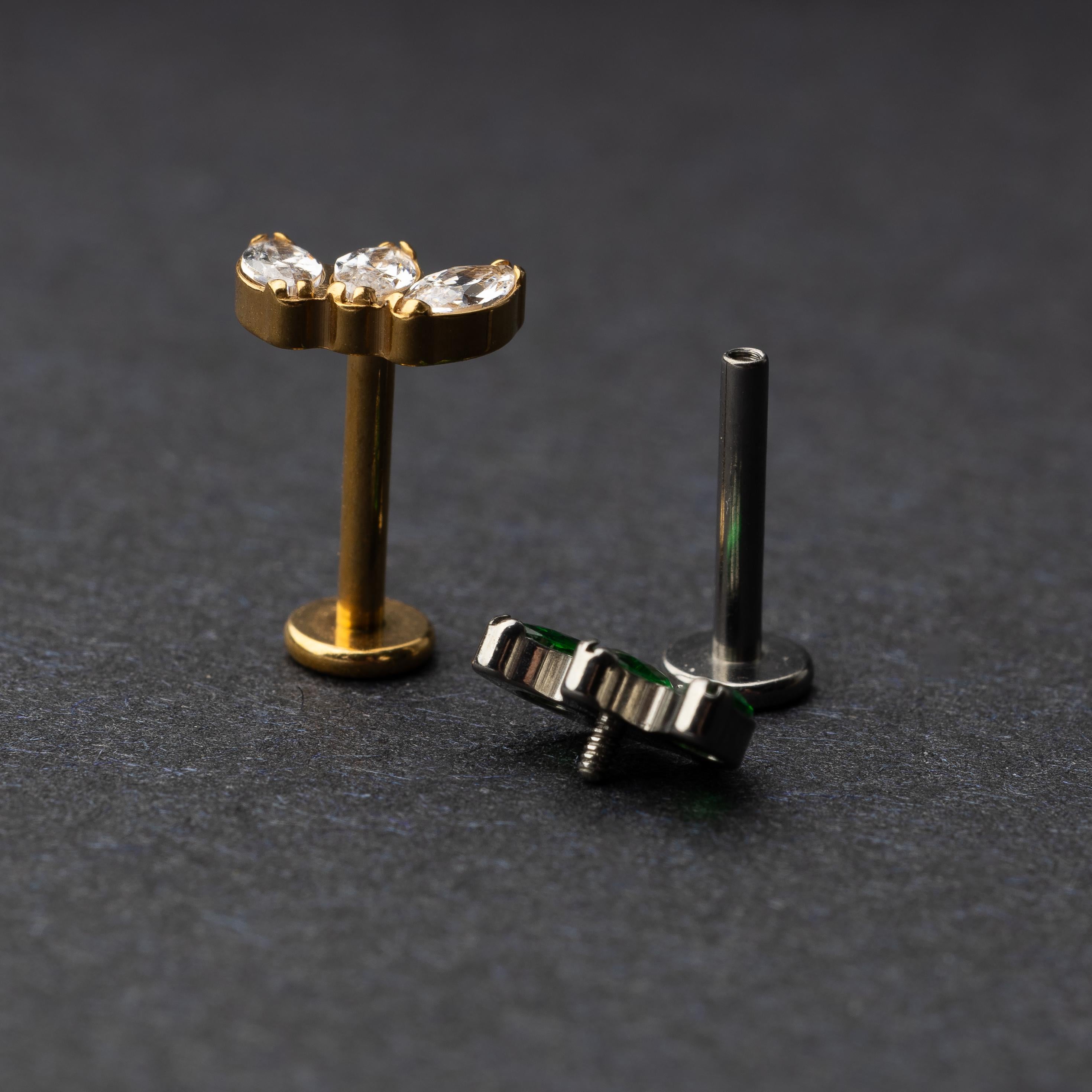 Two flat back bars featuring a triple gem design: one with emerald green gems on a silver base and the other with clear gems on a gold base, displayed on a dark background.