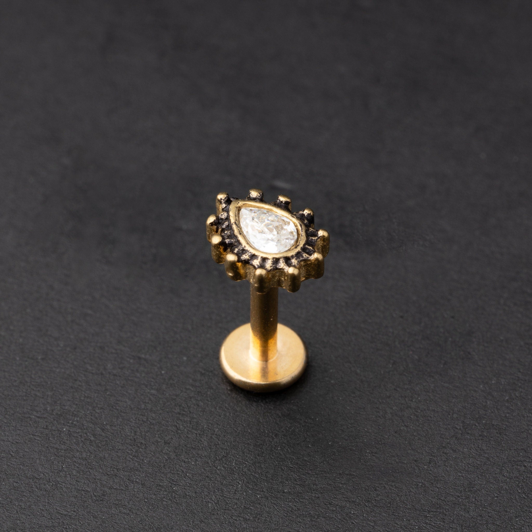 Gold labret stud with a teardrop-shaped crystal centrepiece and detailed border, displayed on a dark background.