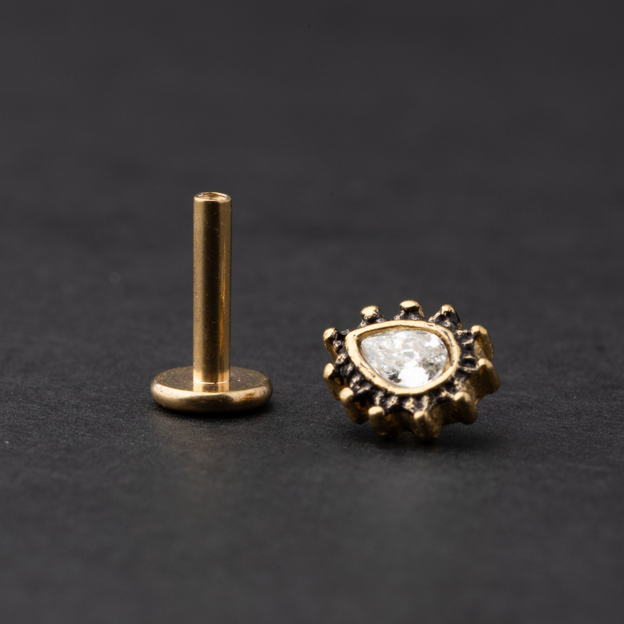 Gold labret stud with a teardrop-shaped crystal centerpiece and detailed border, displayed on a dark background.