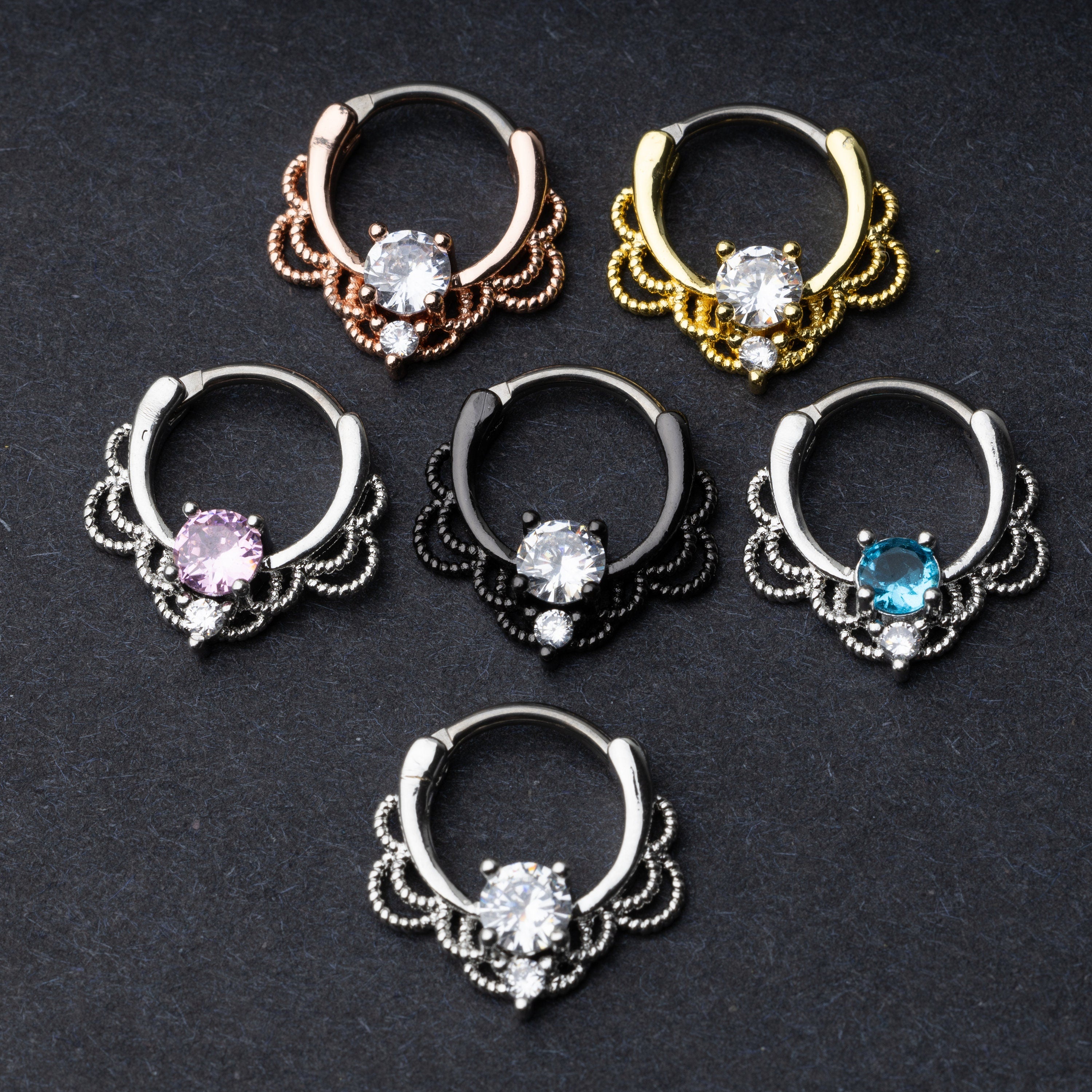 Ornate septum clickers in various finishes (gold, rose gold, black, and silver) with center gemstones in different colors, displayed on a dark background.