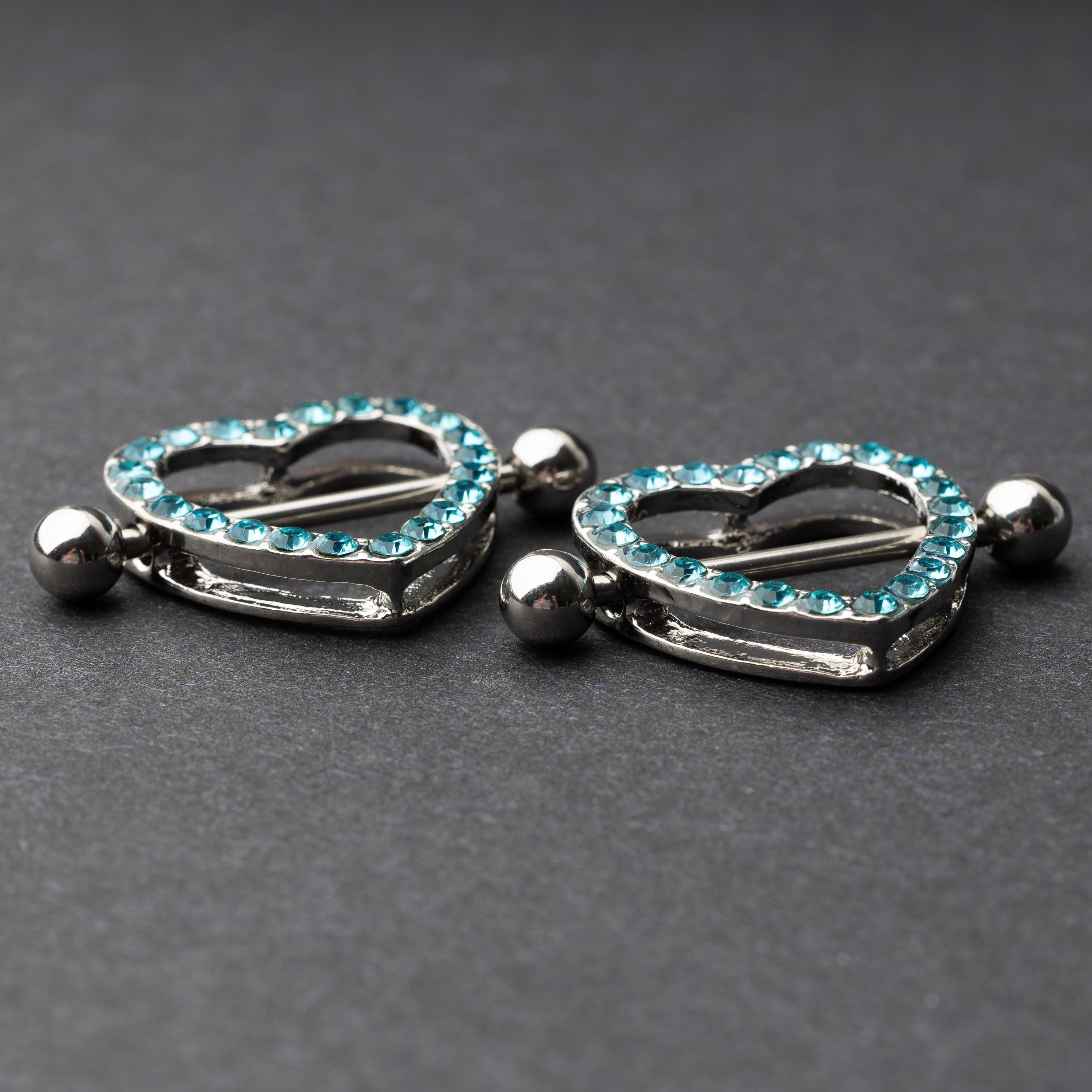 Two heart-shaped nipple barbells with silver metal and light blue gemstone accents, displayed on a dark background.