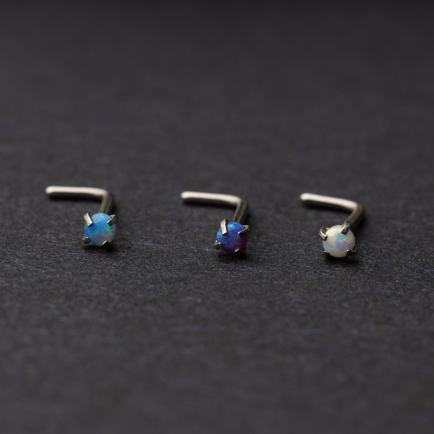 Three L-shaped opalescent nose studs in different shades, displayed on a dark background.