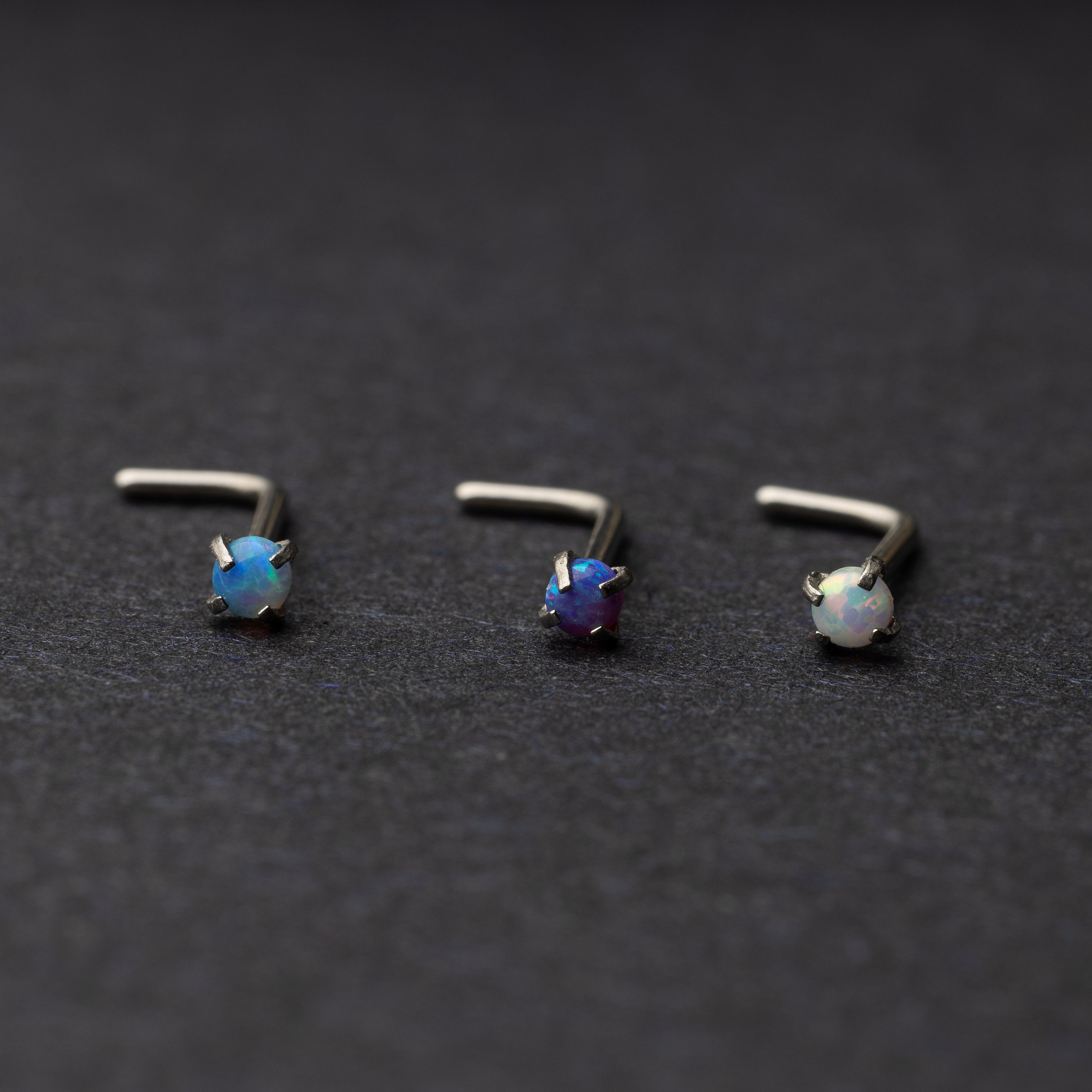 Three L-shaped opalescent nose studs in different shades, displayed on a dark background.