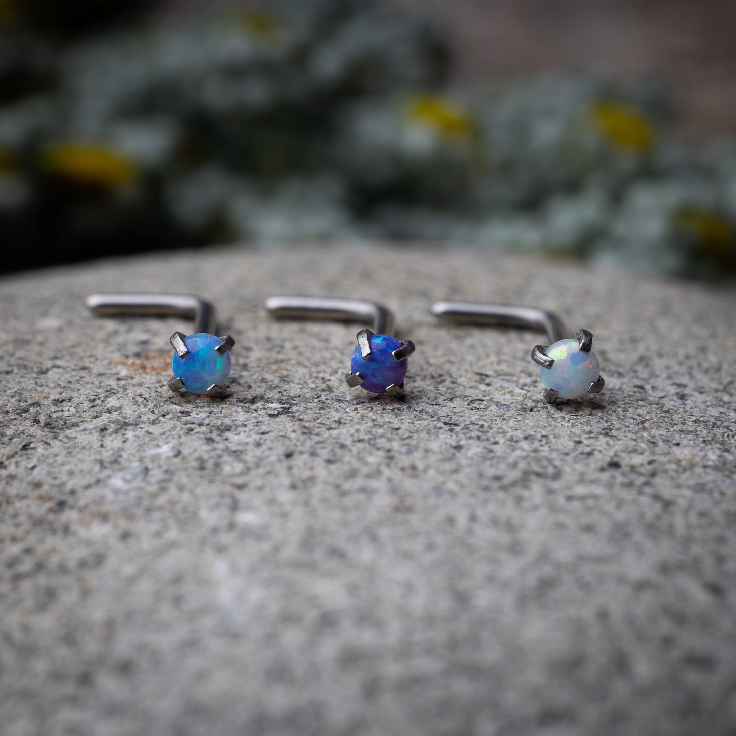 Three L-shaped opalescent nose studs in different shades, displayed on a stone background.