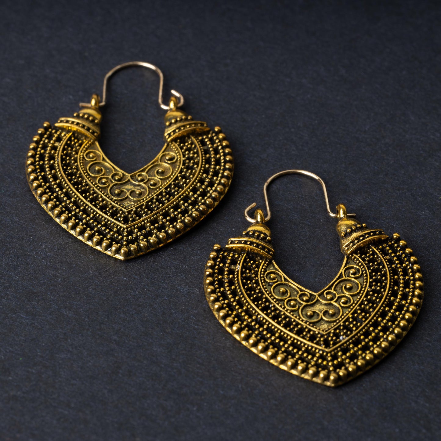 Pair of earrings in antique gold with intricate patterns and textured detailing, displayed on a dark background.