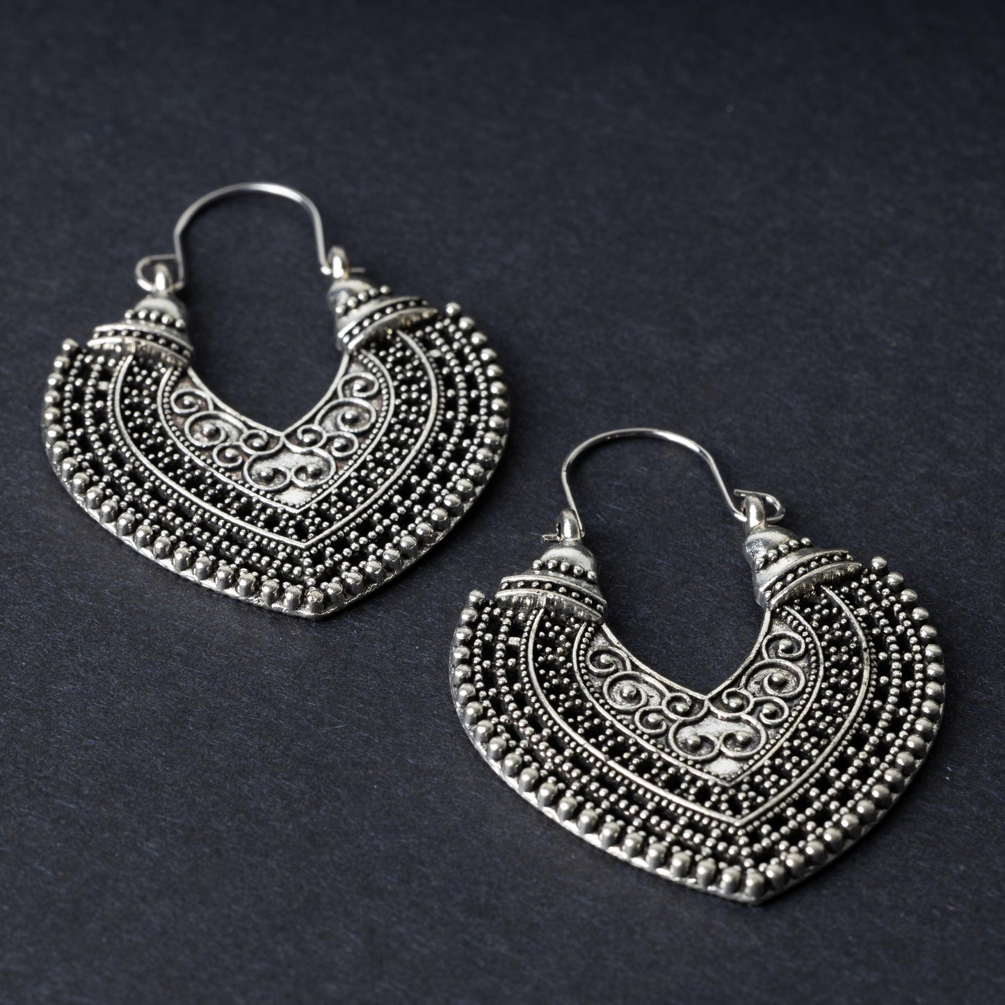 Pair of earrings in antique silver with intricate patterns and textured detailing, displayed on a dark background.