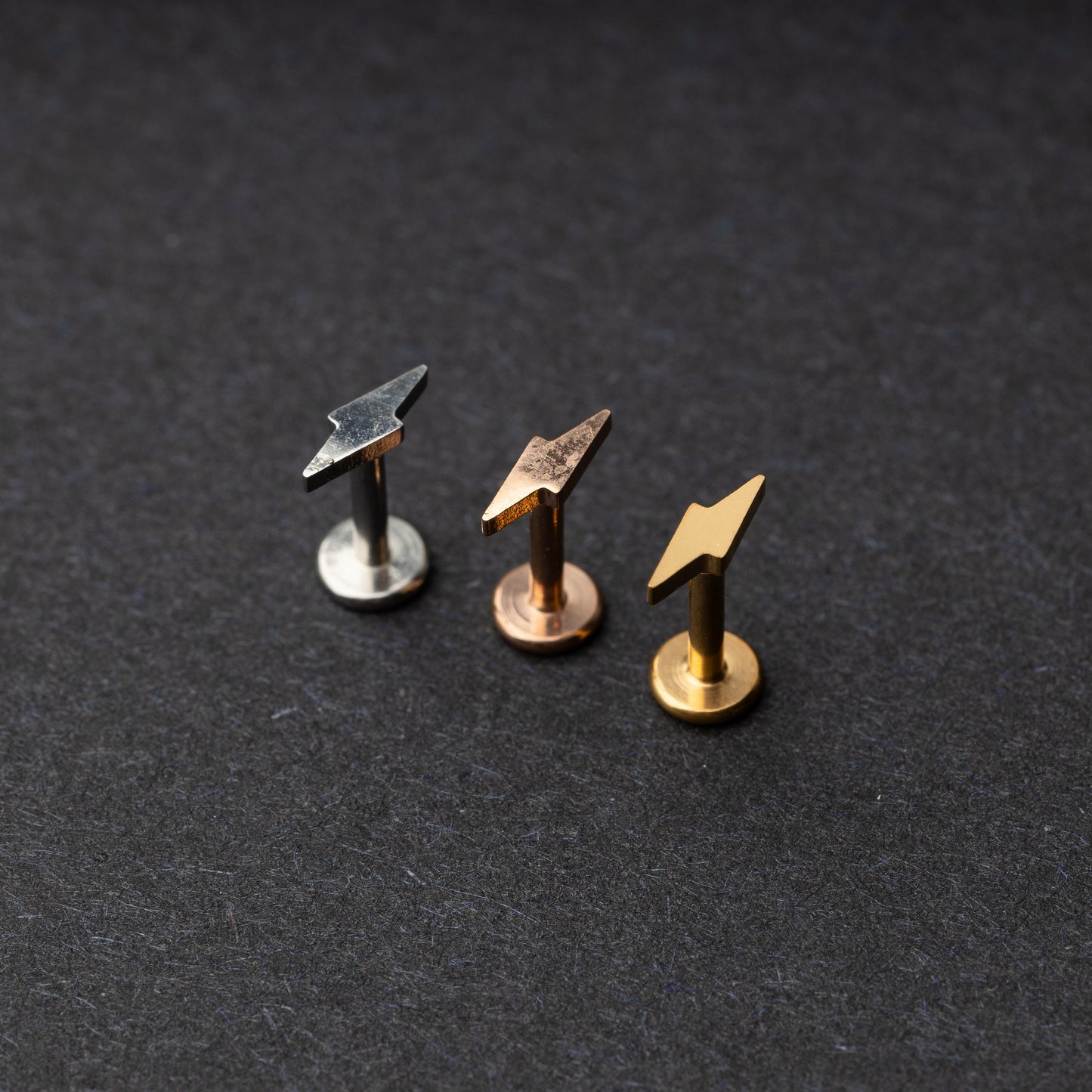 Minimalist lightning bolt labret studs in silver, gold, and rose gold finishes, displayed on a dark background.