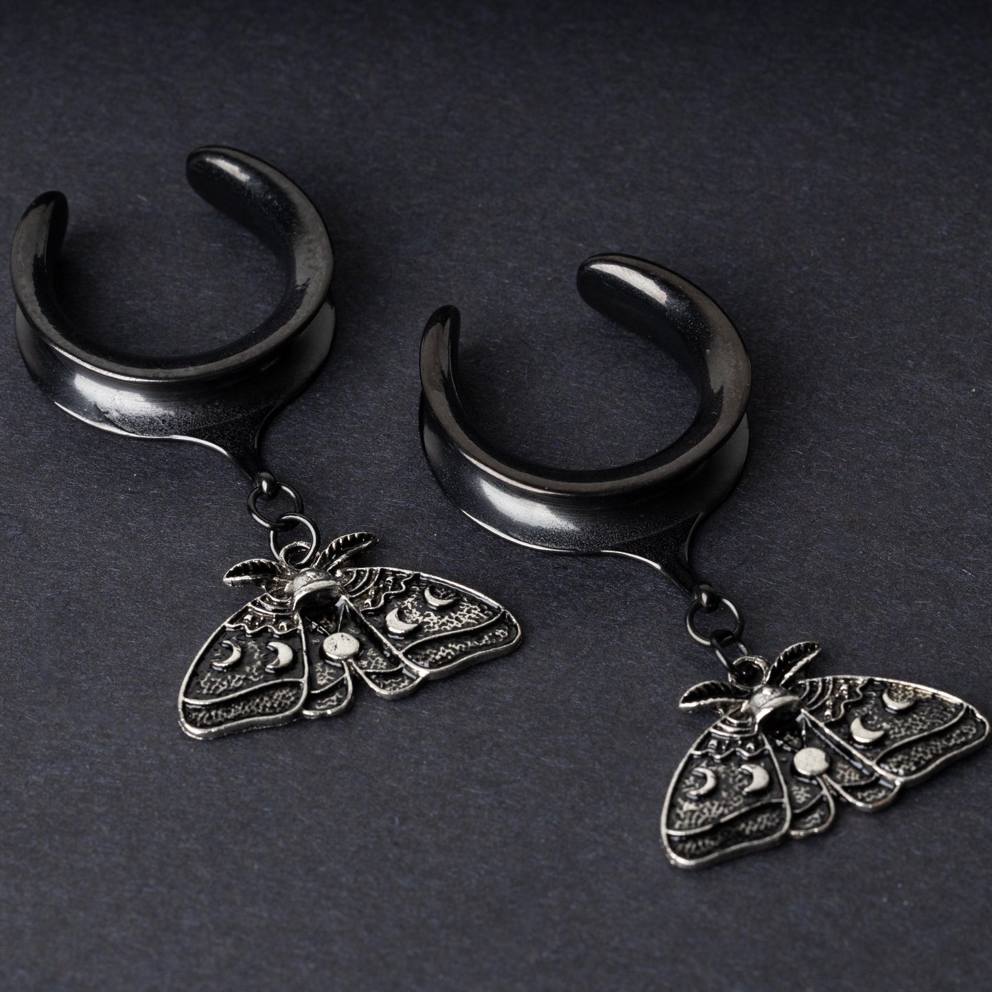 Black tunnels for stretched ears with dangling ornate moth charms featuring celestial patterns, displayed on a dark background.