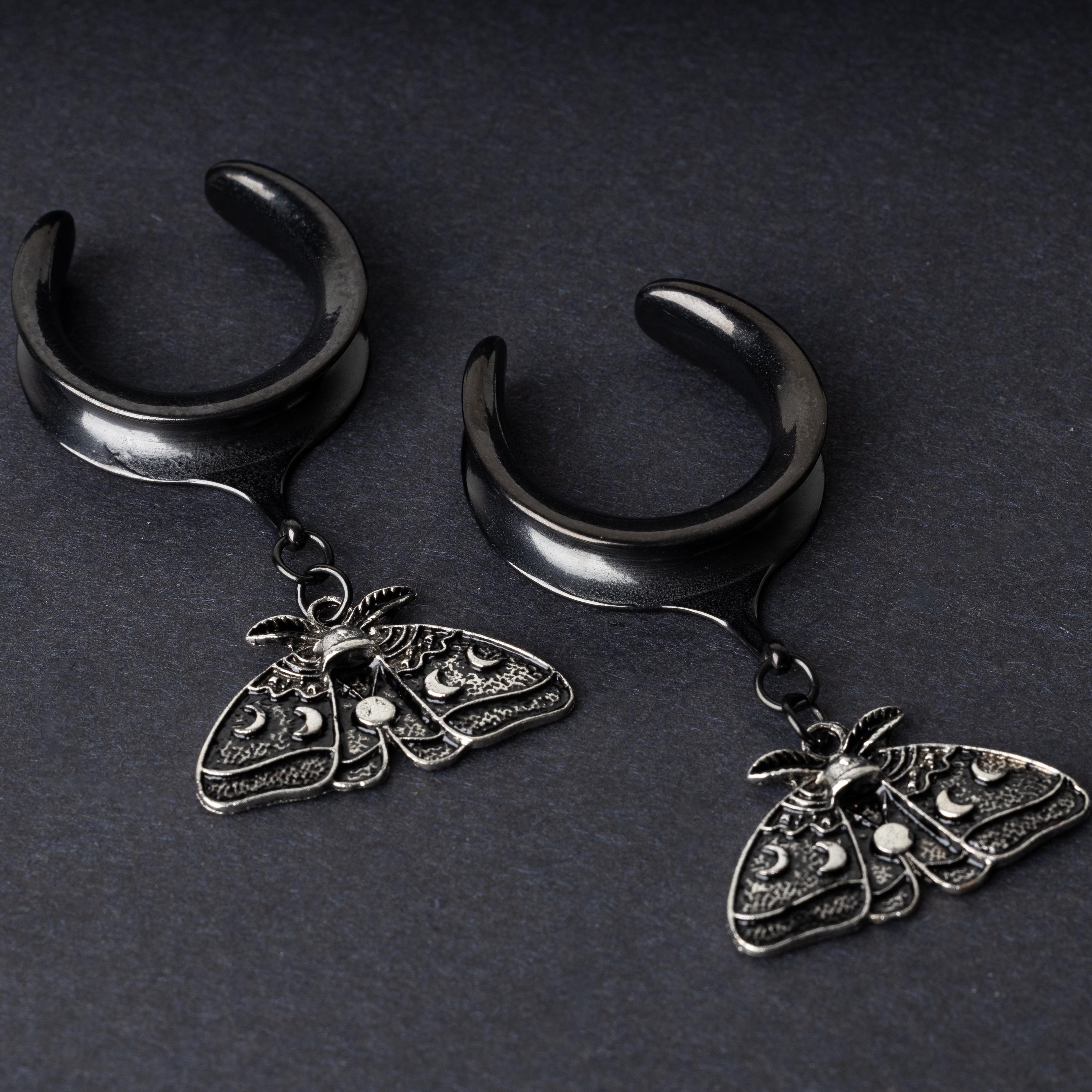 Black tunnels for stretched ears with dangling ornate moth charms featuring celestial patterns, displayed on a dark background.