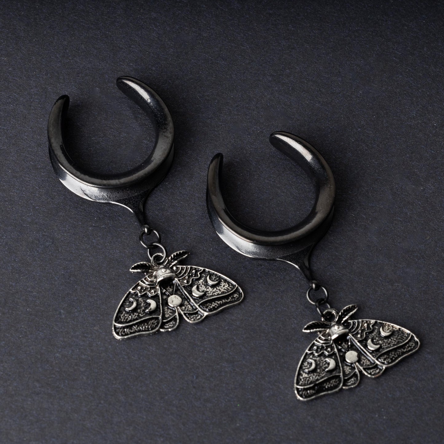 Black tunnels for stretched ears with dangling ornate moth charms featuring celestial patterns, displayed on a dark background.