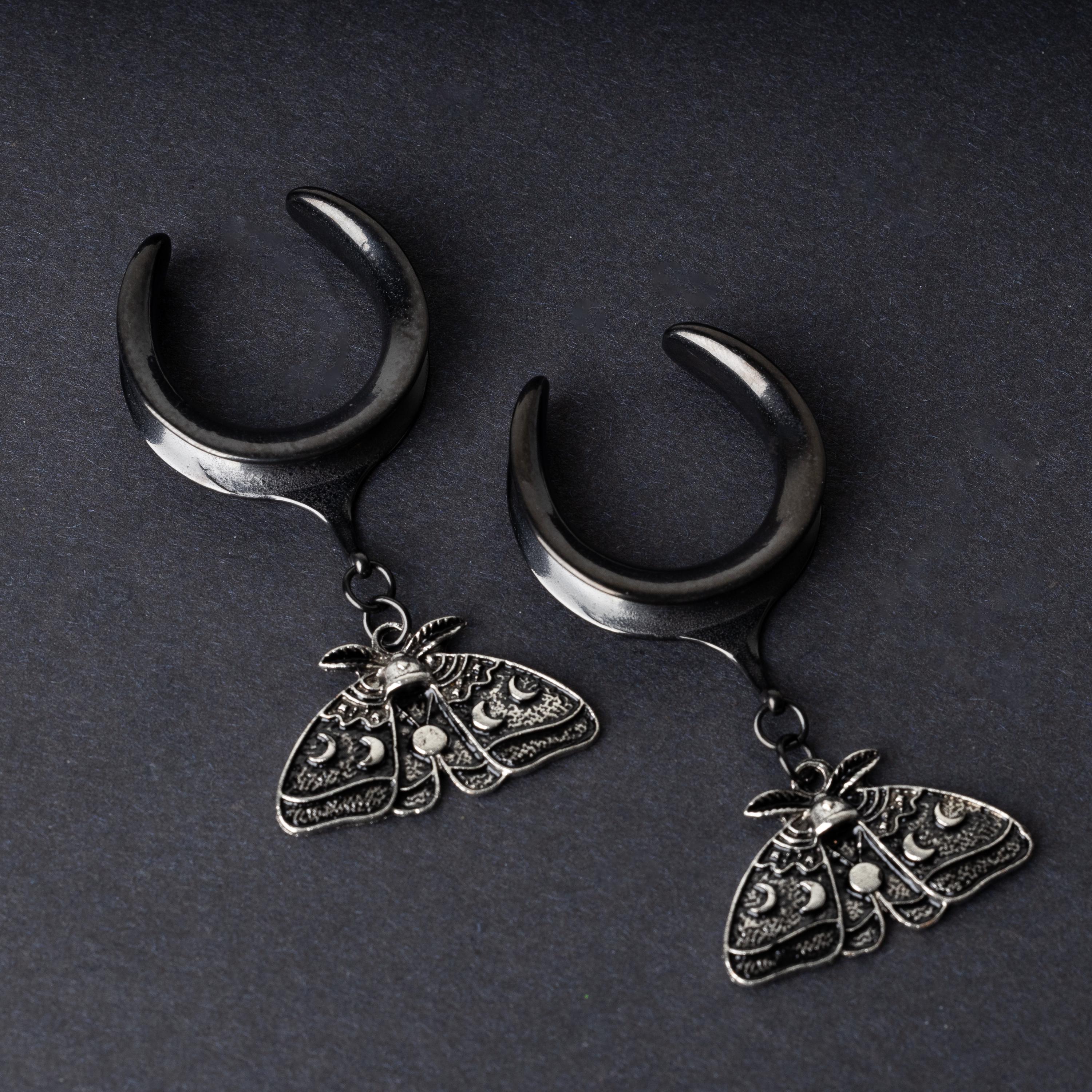 Black tunnels for stretched ears with dangling ornate moth charms featuring celestial patterns, displayed on a dark background.