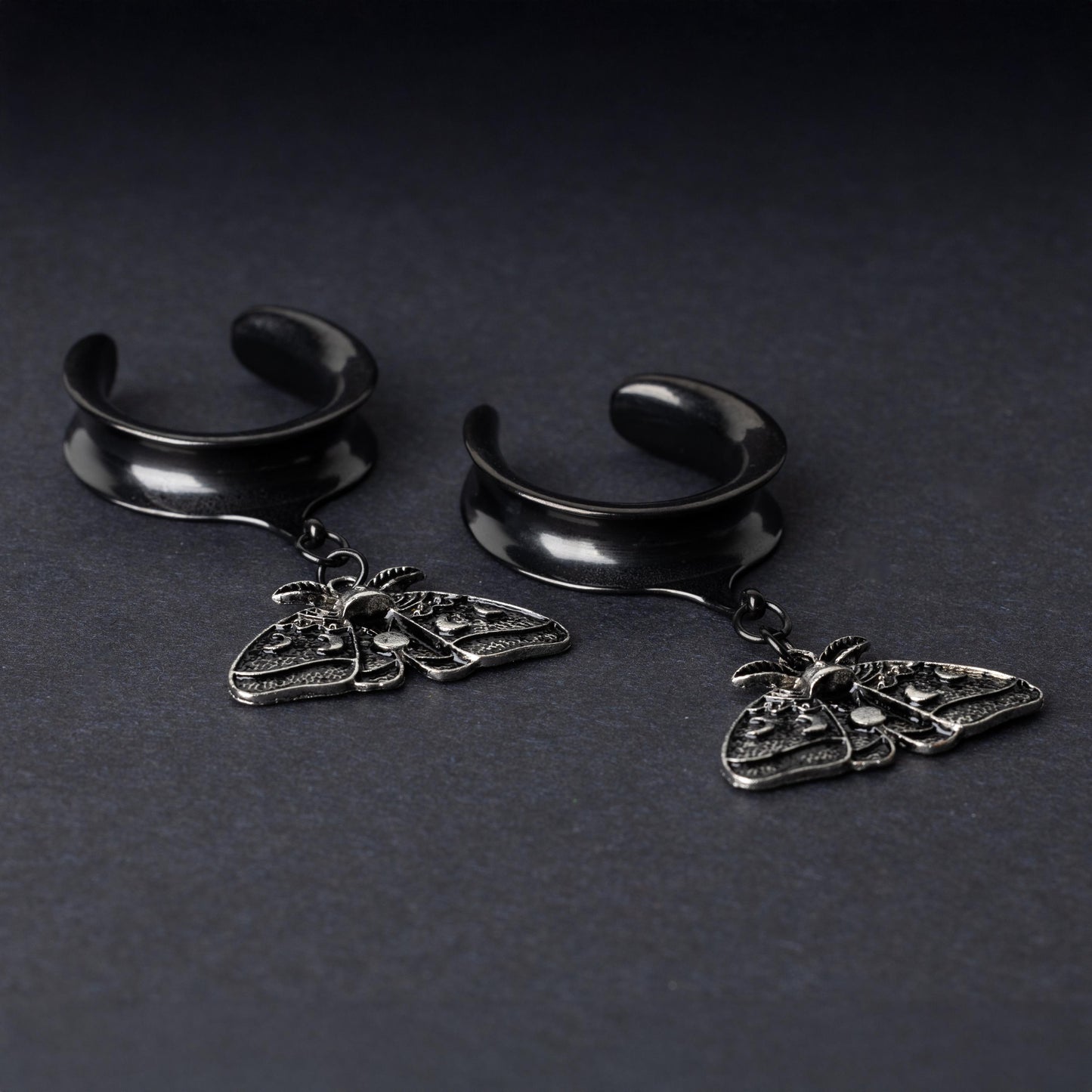 Black tunnels for stretched ears with dangling ornate moth charms featuring celestial patterns, displayed on a dark background.