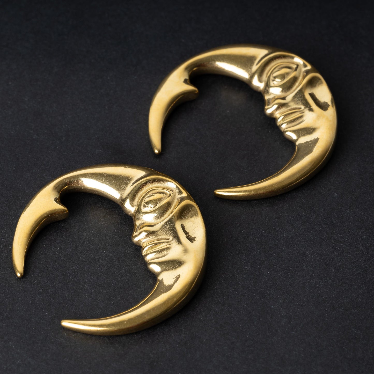 A pair of crescent moon-shaped ear weights in polished gold, featuring a detailed moon face design, displayed on a dark background.