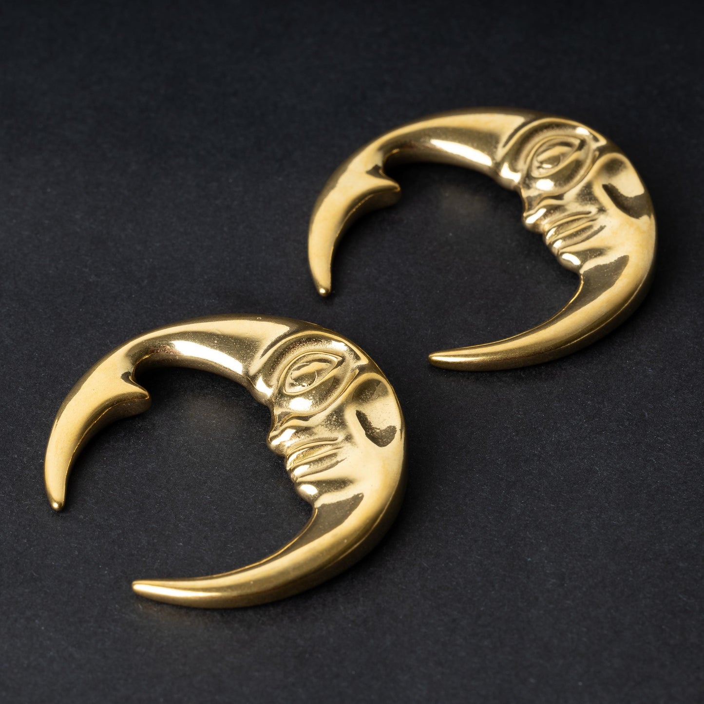 A pair of crescent moon-shaped ear weights in polished gold, featuring a detailed moon face design, displayed on a dark background.