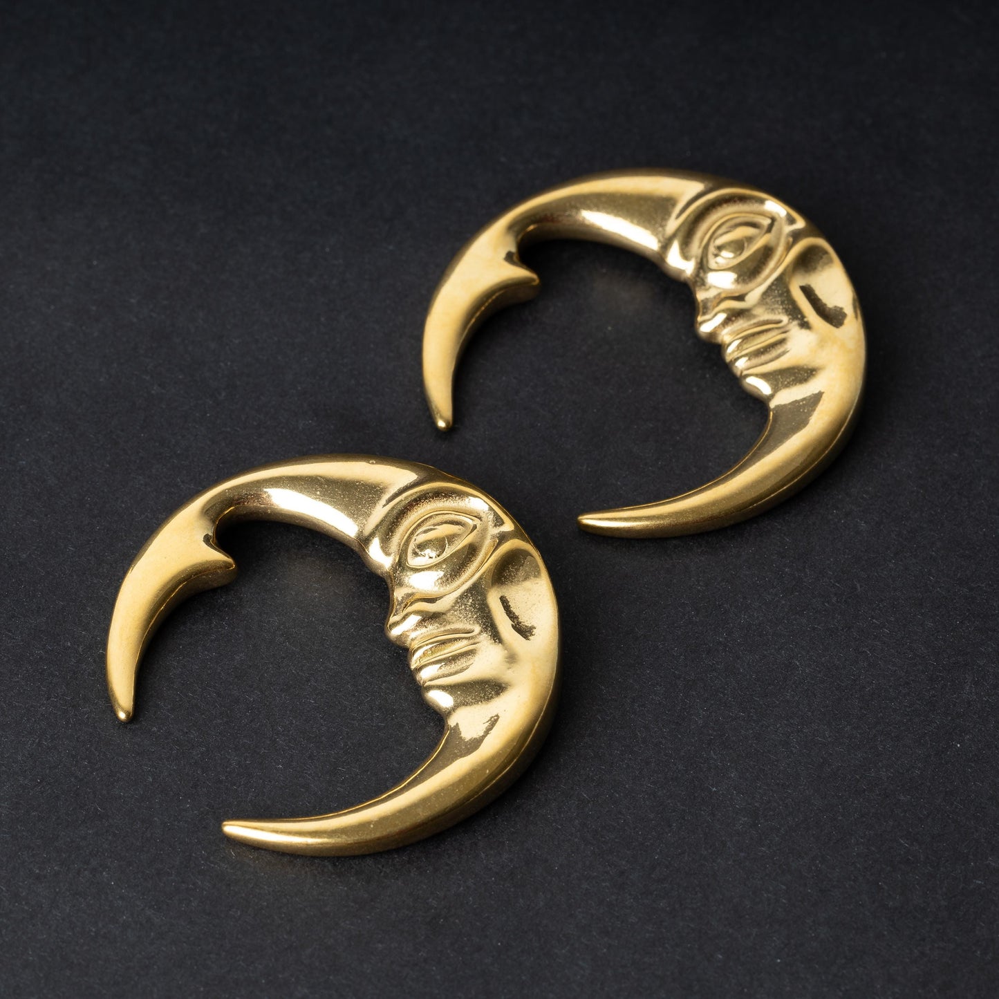 A pair of crescent moon-shaped ear weights in polished gold, featuring a detailed moon face design, displayed on a dark background.