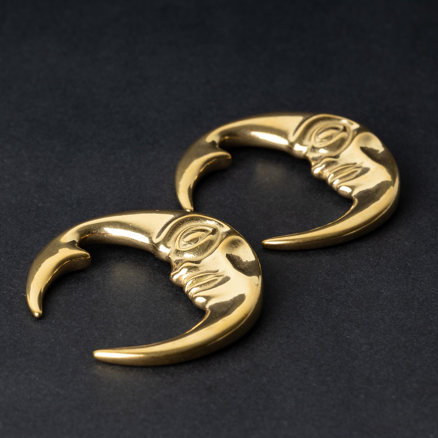 A pair of crescent moon-shaped ear weights in polished gold, featuring a detailed moon face design, displayed on a dark background.