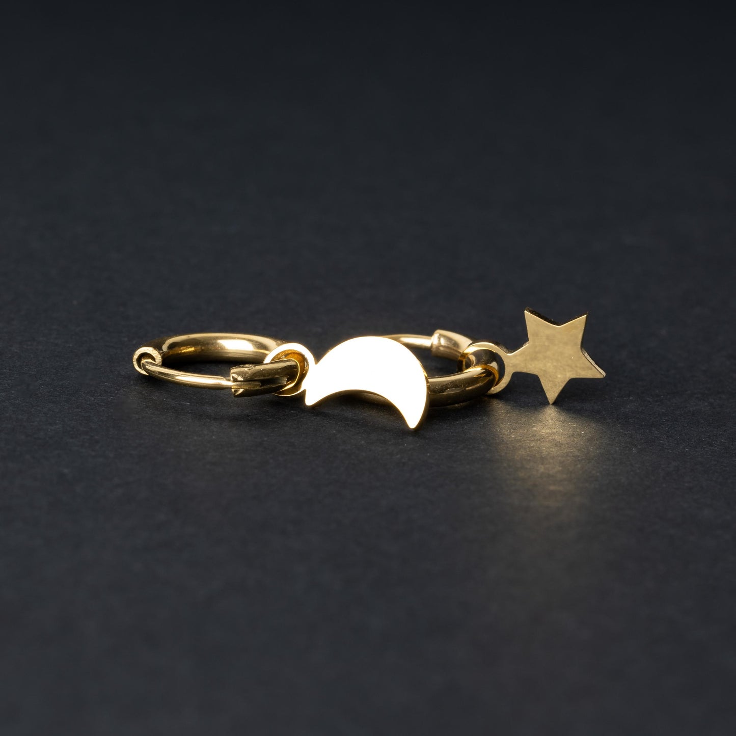 Gold huggie hoop earrings featuring a crescent moon charm on one hoop and a star charm on the other, displayed on a dark background.